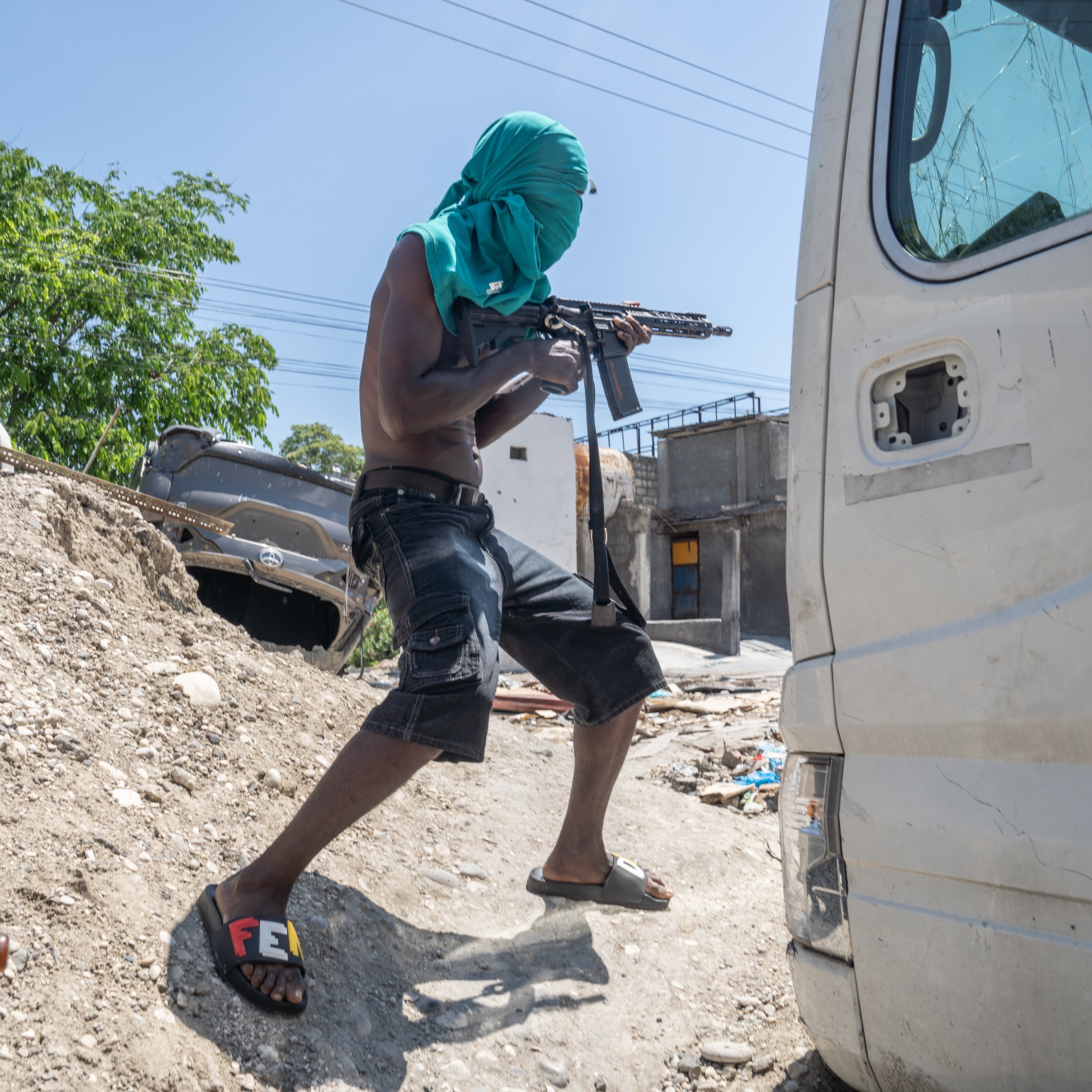 A person with their face covered with a turquoise t-shirt is holding a rifle, crouching beside a damaged vehicle in an urban setting with debris and damaged buildings in the background.