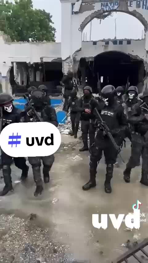 A group of heavily armed individuals dressed in tactical gear and helmets stands in front of a partially demolished building, with signs of destruction visible in the surroundings.
