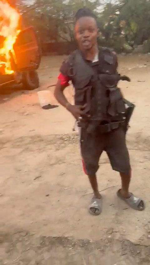 A person stands on a dirt road wearing a tactical vest and shorts, with a large fire blazing in a vehicle behind them.
