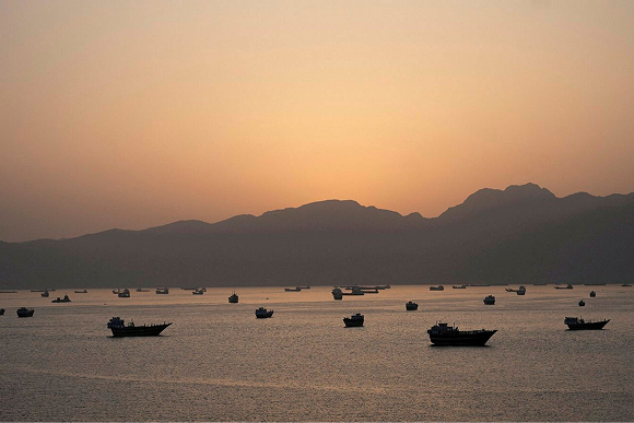 Alt text 1: Vessels in silhouette scattered across calm water in the Strait of Hormuz at sunset with mountains in the background.