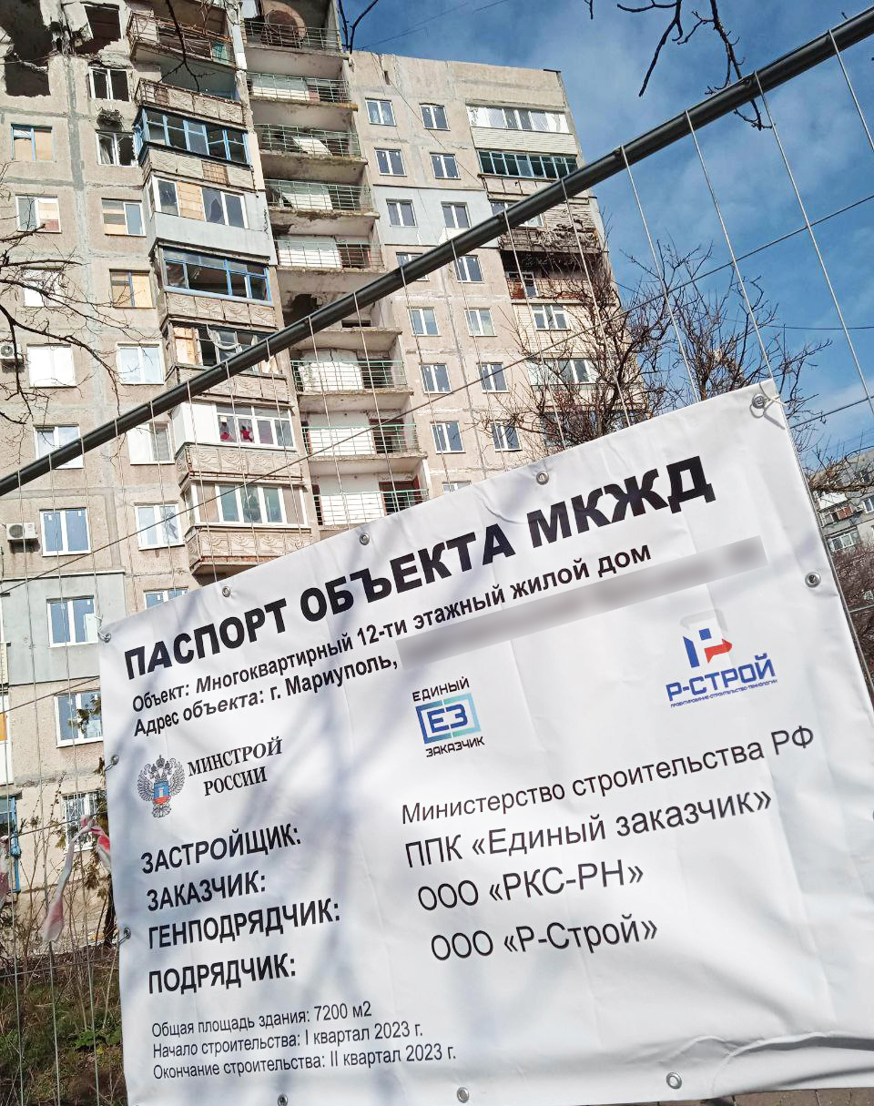 A large poster hung on the fence of a construction site, with text in Russian describing the project and those responsible. An arrow points to a section, which is translated as: