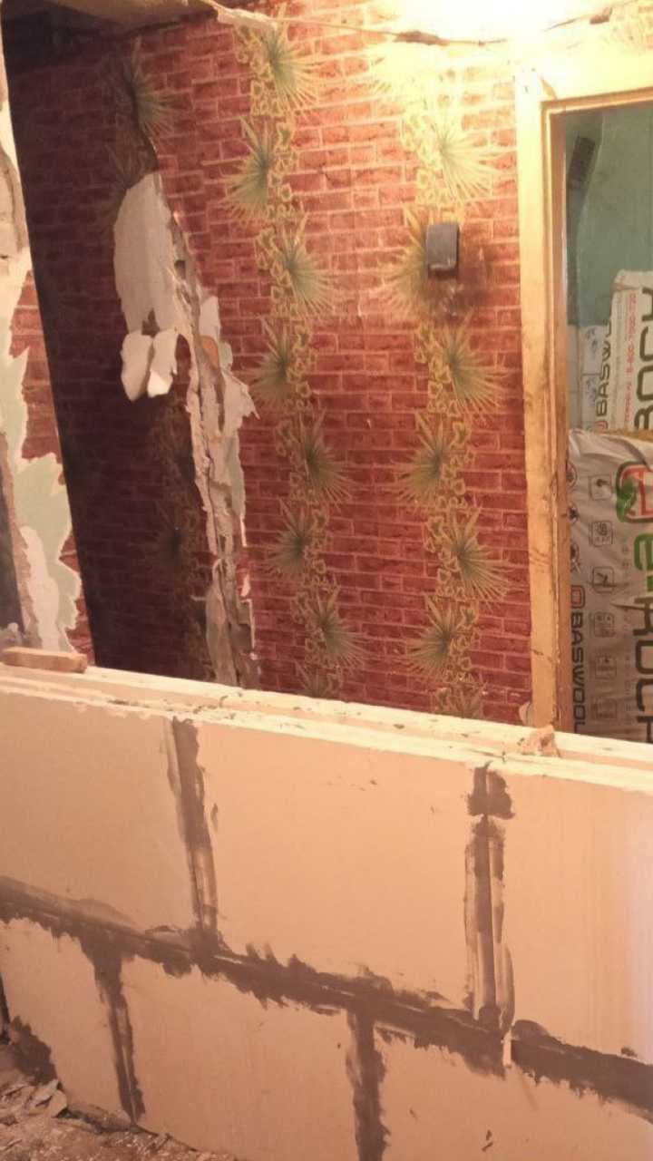 Still image from inside an apartment, showing a half-built cinderblock wall where a previous interior wall had collapsed. Over the waist-high wall, the apartment next door is visible.