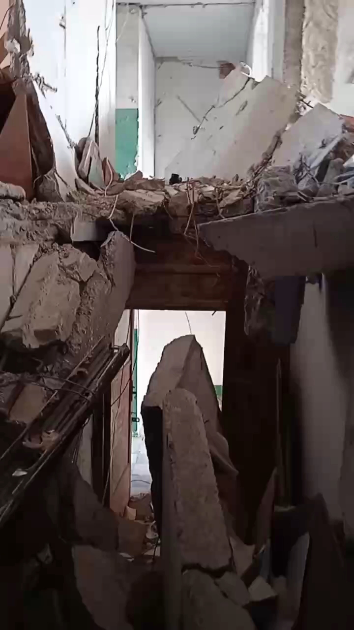 Still image from video of a damaged hallway inside an apartment building. There is a large hole in the floor, filled with rubble.