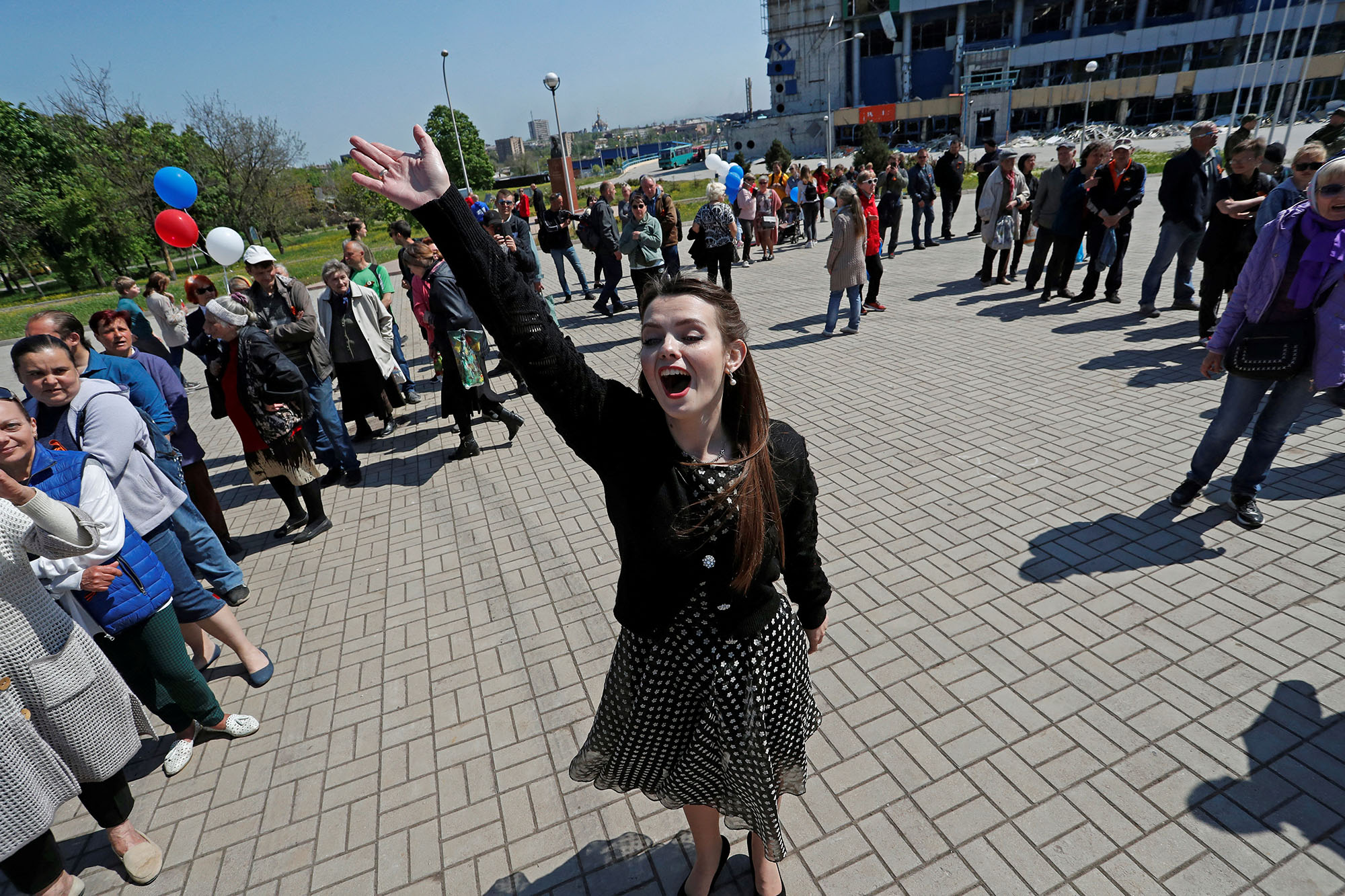 A woman sings in the centre of a large circle of people standing in a city square in Mariupol, celebrating the victory day holiday. Some hold balloons in the blue, white, and red colours of the Russian flag.