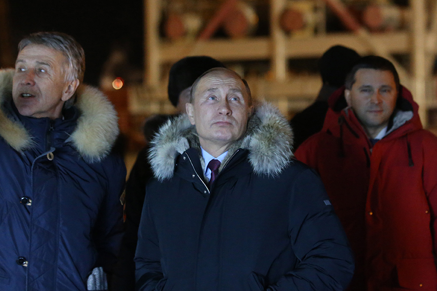 La nuit, trois hommes se tiennent devant une grande tour en acier, vêtus de lourdes parkas.  Un homme à gauche montre quelque chose derrière la caméra, tandis que Vladimir Poutine se tient au milieu, regardant avec une expression d'intérêt.