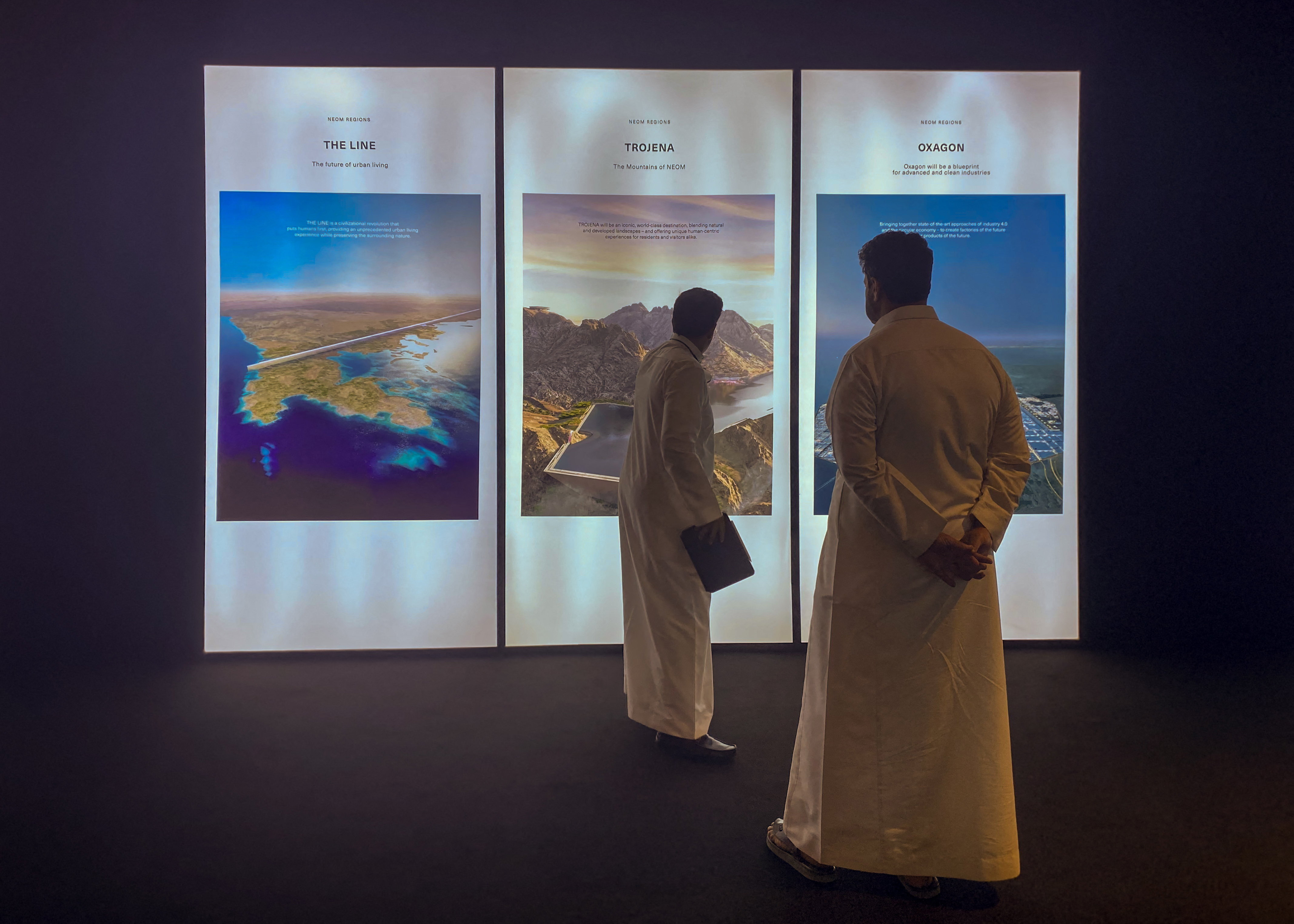 Two men in white robes view three backlit posters labeled "THE LINE", "TROJENA" and "OXAGON" showing aerial renderings of planned developments.