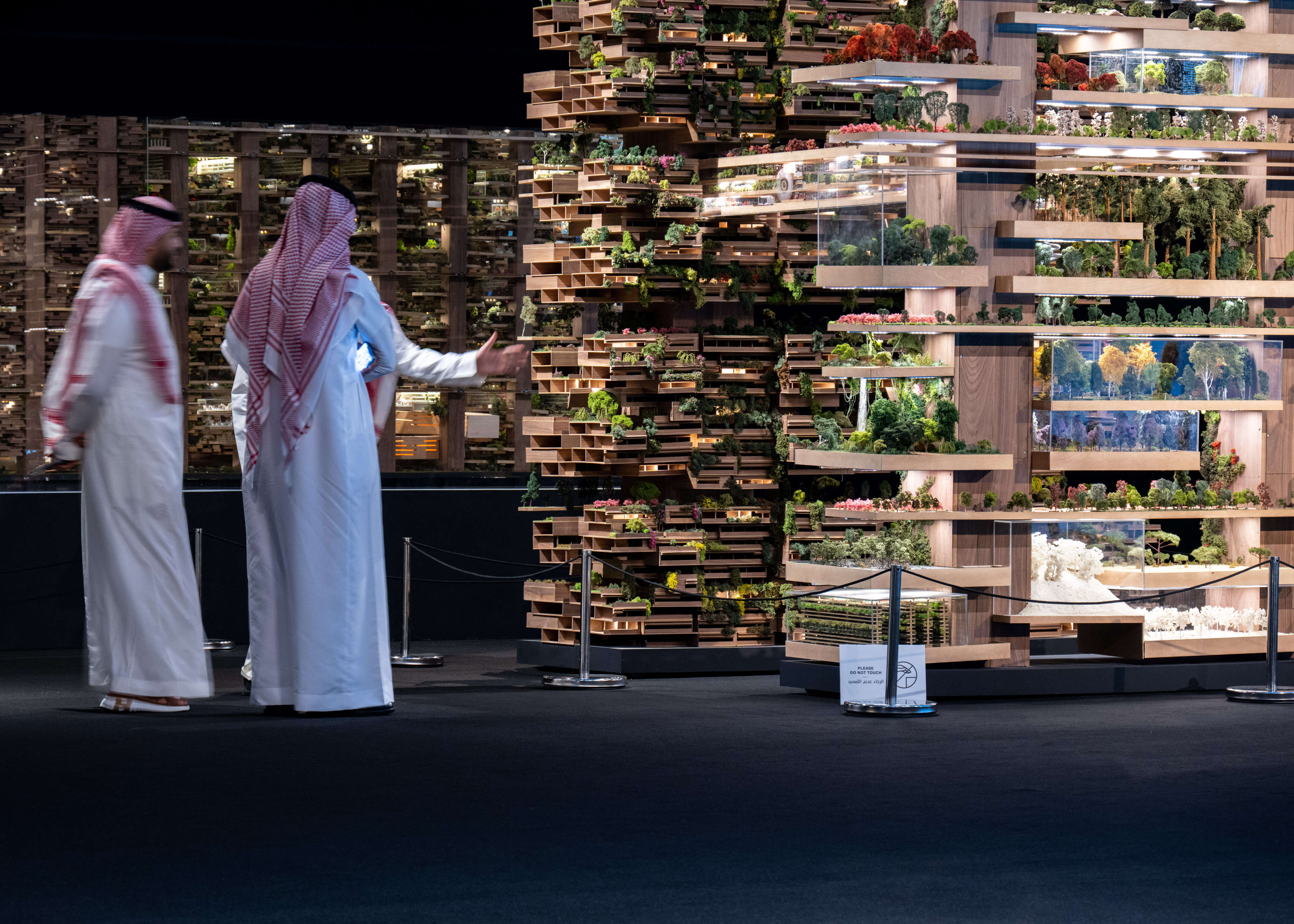 Two men in white thobes and red‑checked keffiyehs gesture toward a large layered architectural model of terraced platforms filled with miniature trees and gardens.