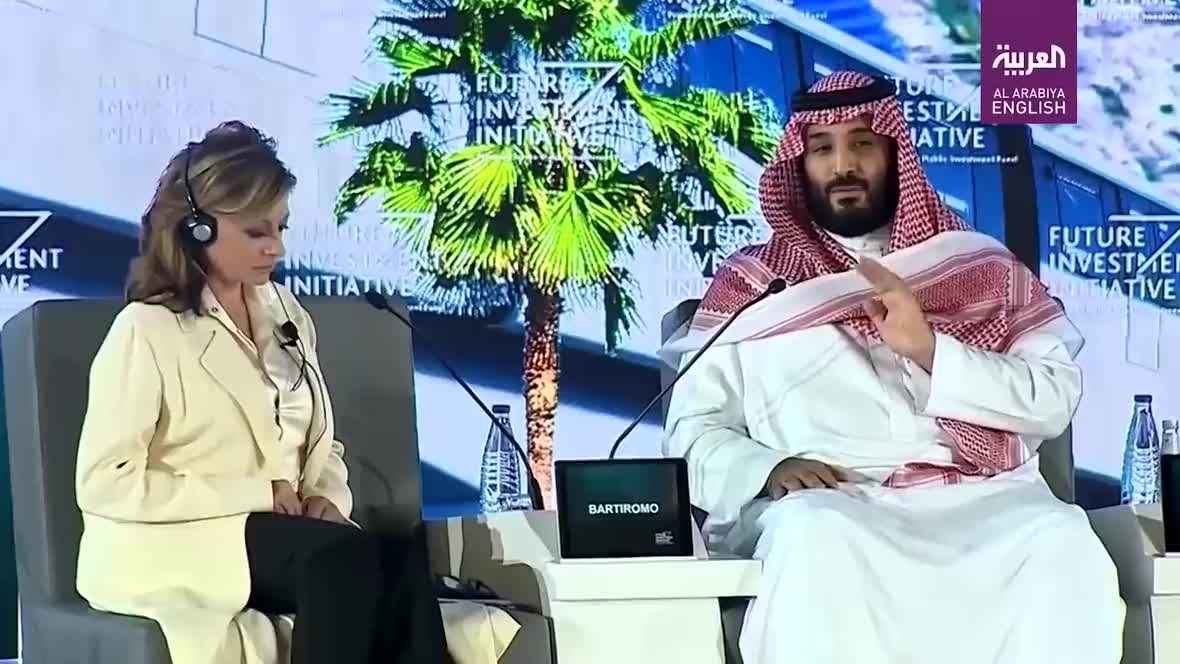 Man in white thobe and red headscarf beside a woman with a headset and light blazer on a stage, 'Future Investment Initiative' logo visible behind them.