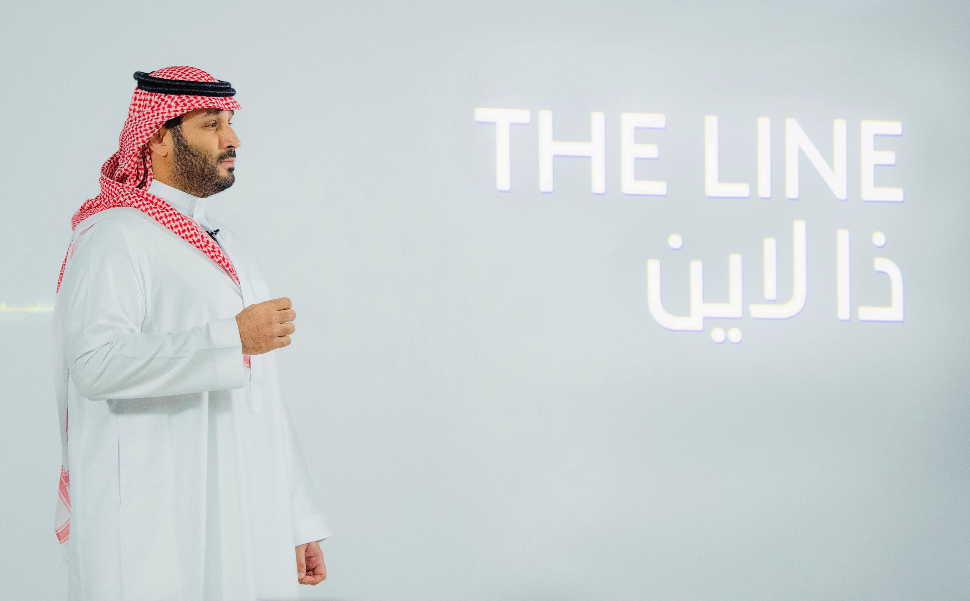 A man in a white thobe and red-and-white keffiyeh stands left, looking toward projected text "THE LINE" with Arabic below.