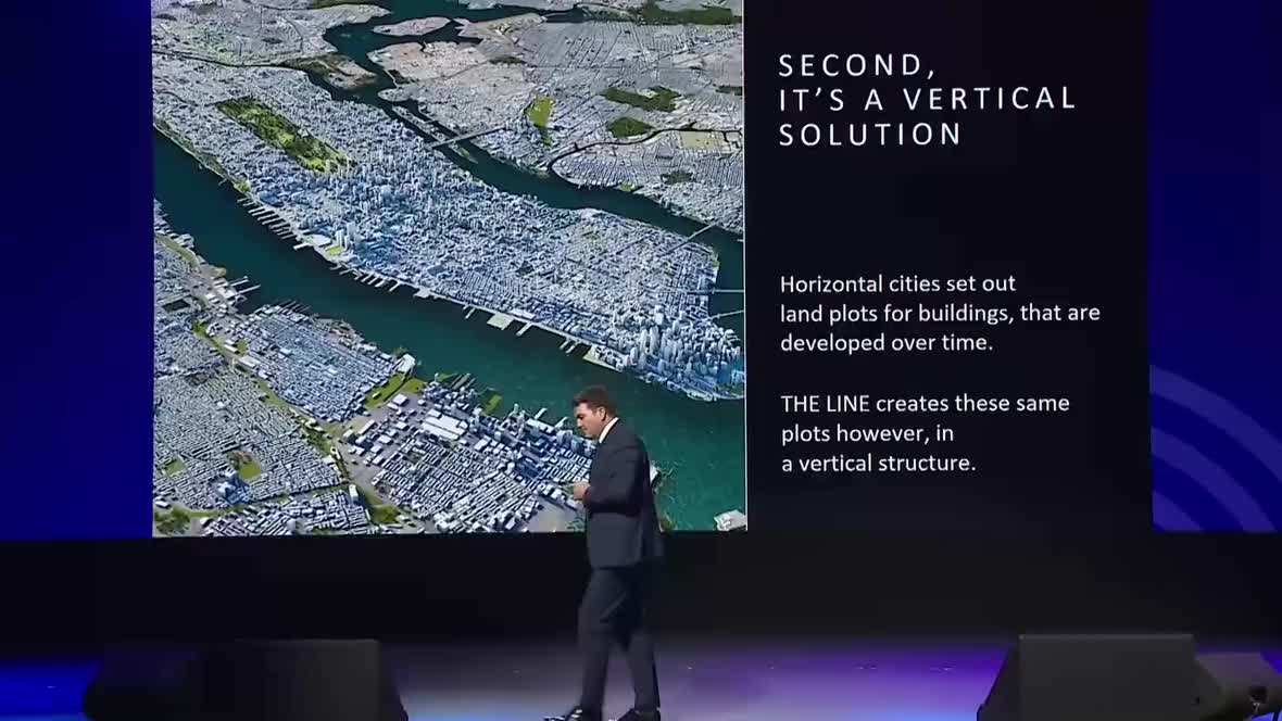 A suited presenter walks across a stage in front of a large screen showing an aerial city map 