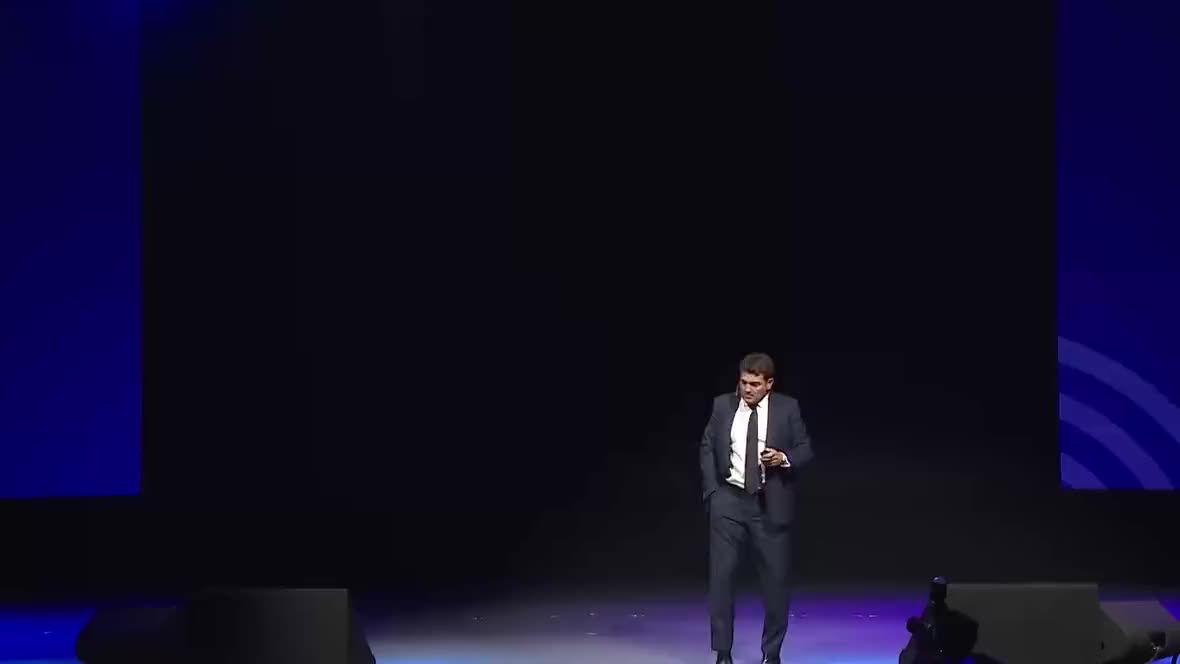 A man in a navy suit and tie stands alone on a large, dimly lit stage holding a microphone under a spotlight.