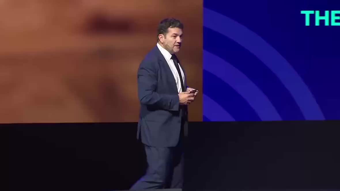 Man in a navy suit speaking on stage, holding a remote and wearing a headset mic. On-screen caption reads, "So we're busy on site as we speak."