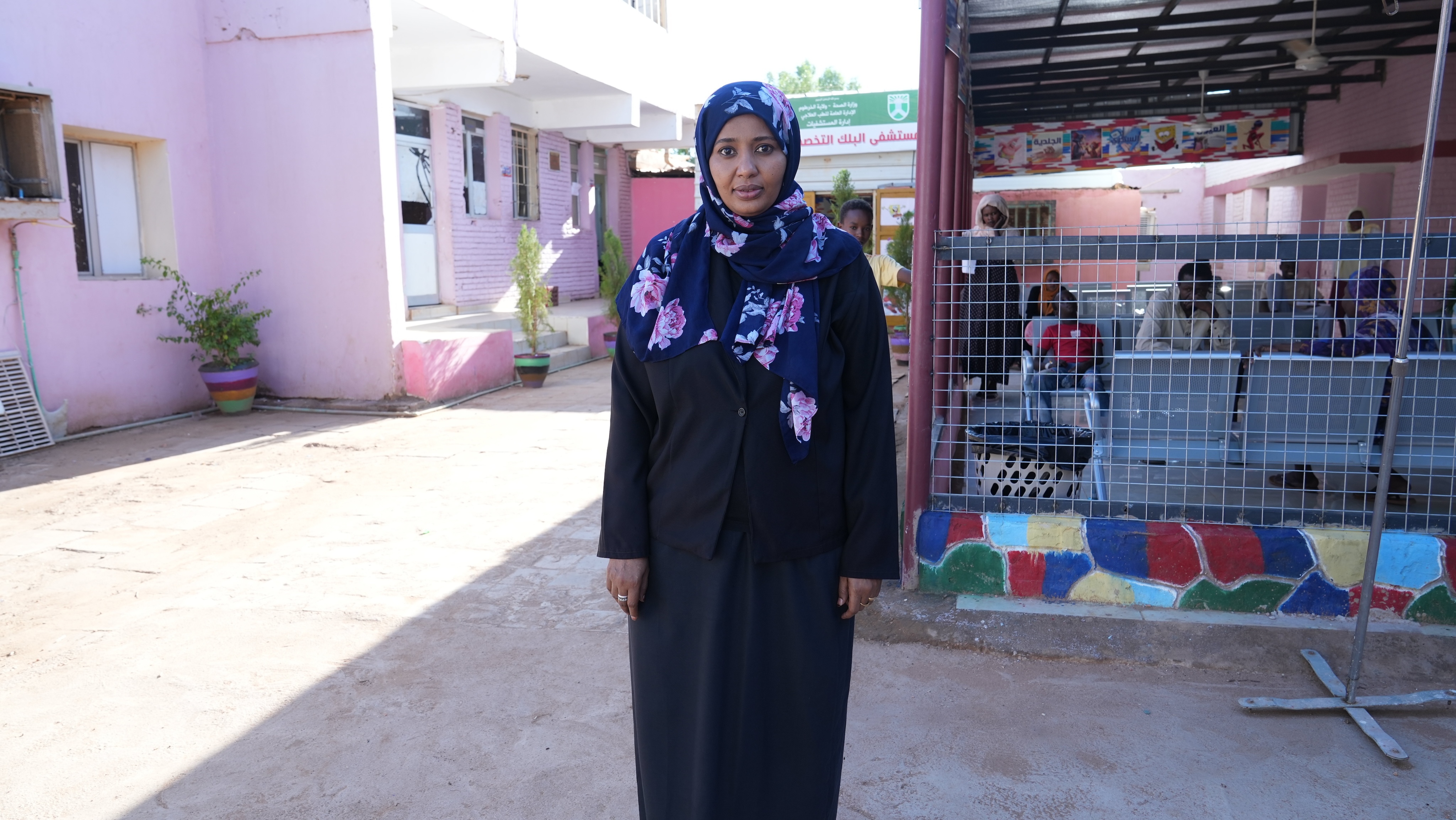 A woman wearing a dark blue dress and floral hijab stands in the courtyard area outside a pink building