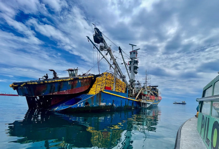 Alt text 1: A brightly-coloured tuna fishing vessel docked beside another vessel on calm blue water under a cloudy sky.