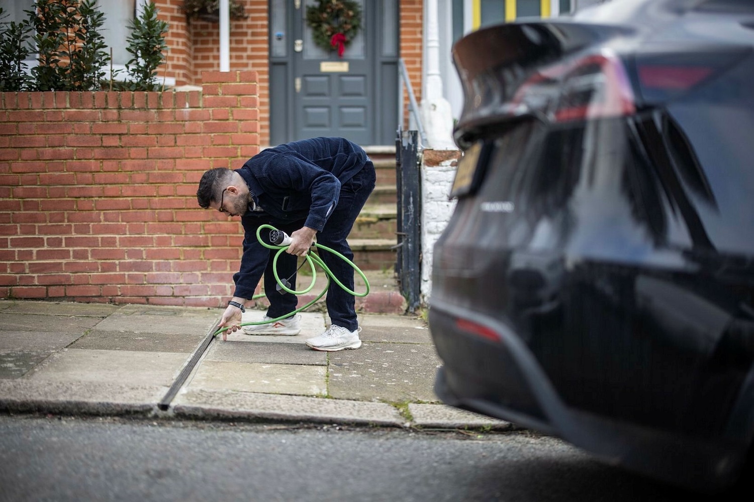 EV owner Chris Richmond, dressed in a dark blue shirt and trousers, places his green charging cable into a narrow gully below a rubber cover outside his terraced property