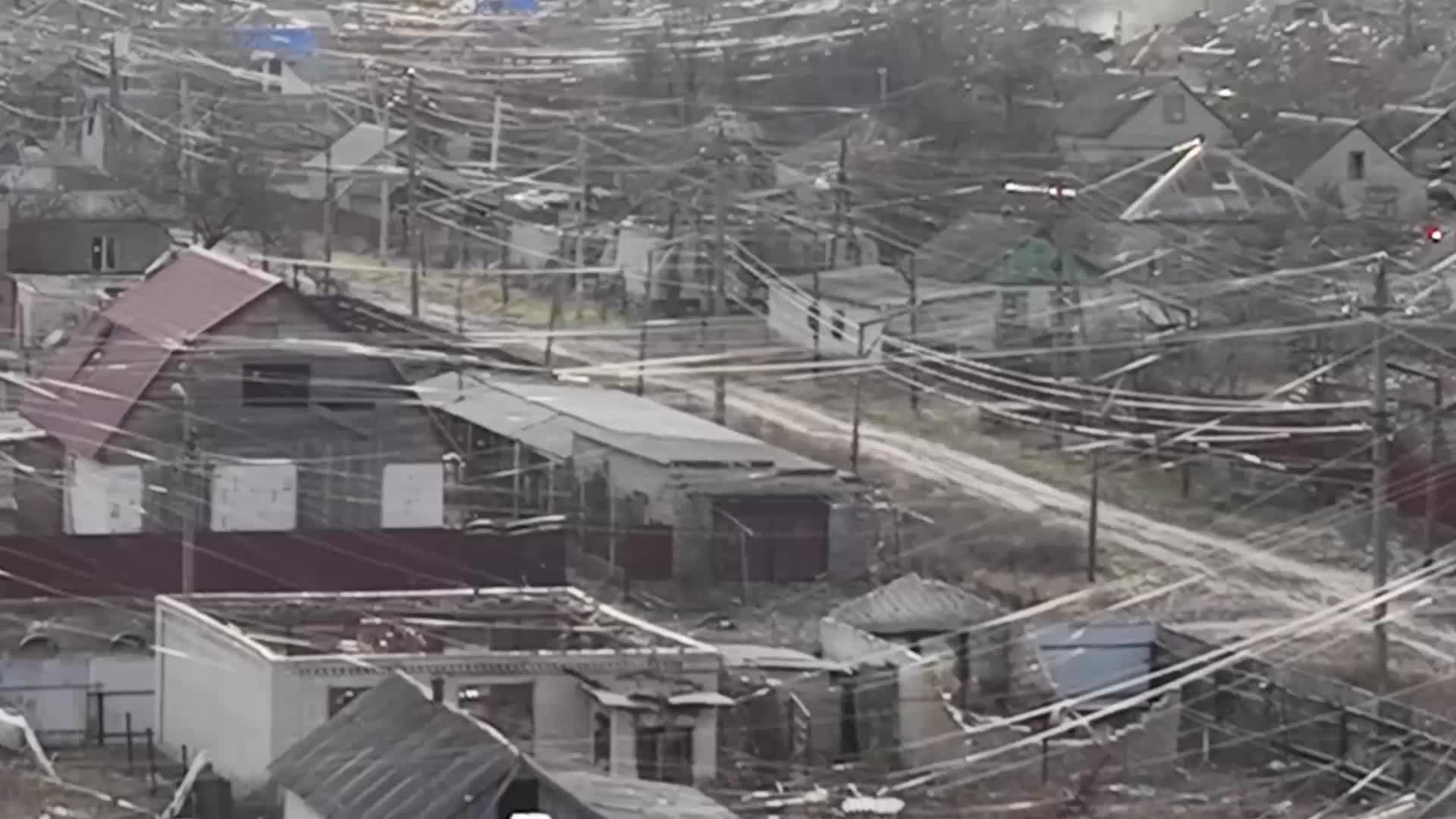 Dense network of overhead utility wires crisscrossing above a cluster of small buildings and a narrow unpaved road.
