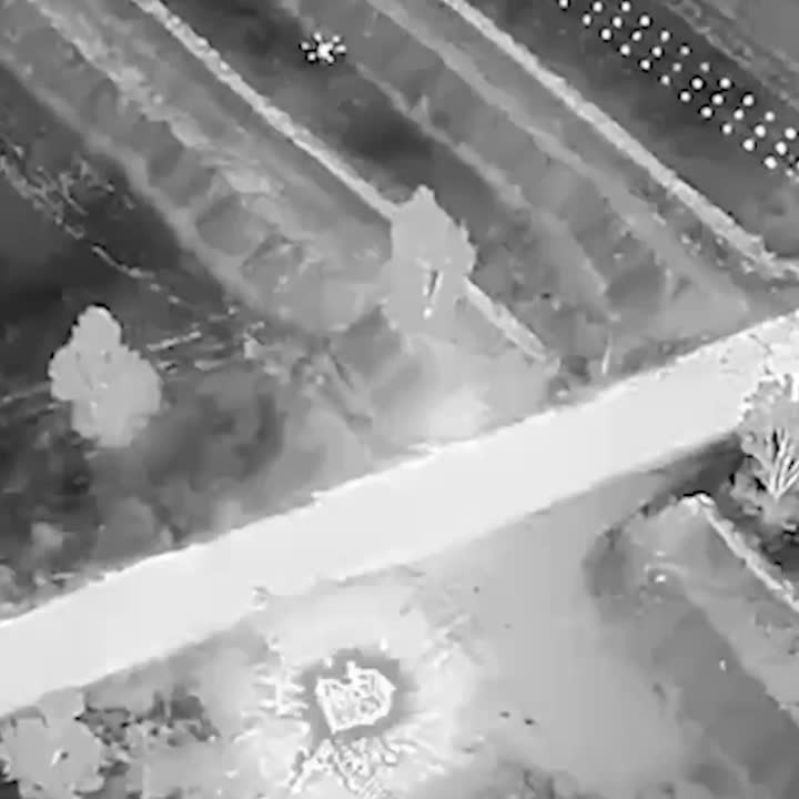 Aerial thermal-style view showing a bright strike along a diagonal road, a fiery explosion and crater beside farmland plots.