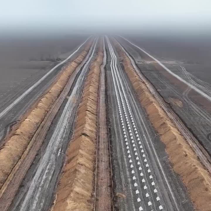 Aerial view of parallel trenches and earthen mounds in a barren field, one trench marked by white triangular markers receding into a foggy horizon.