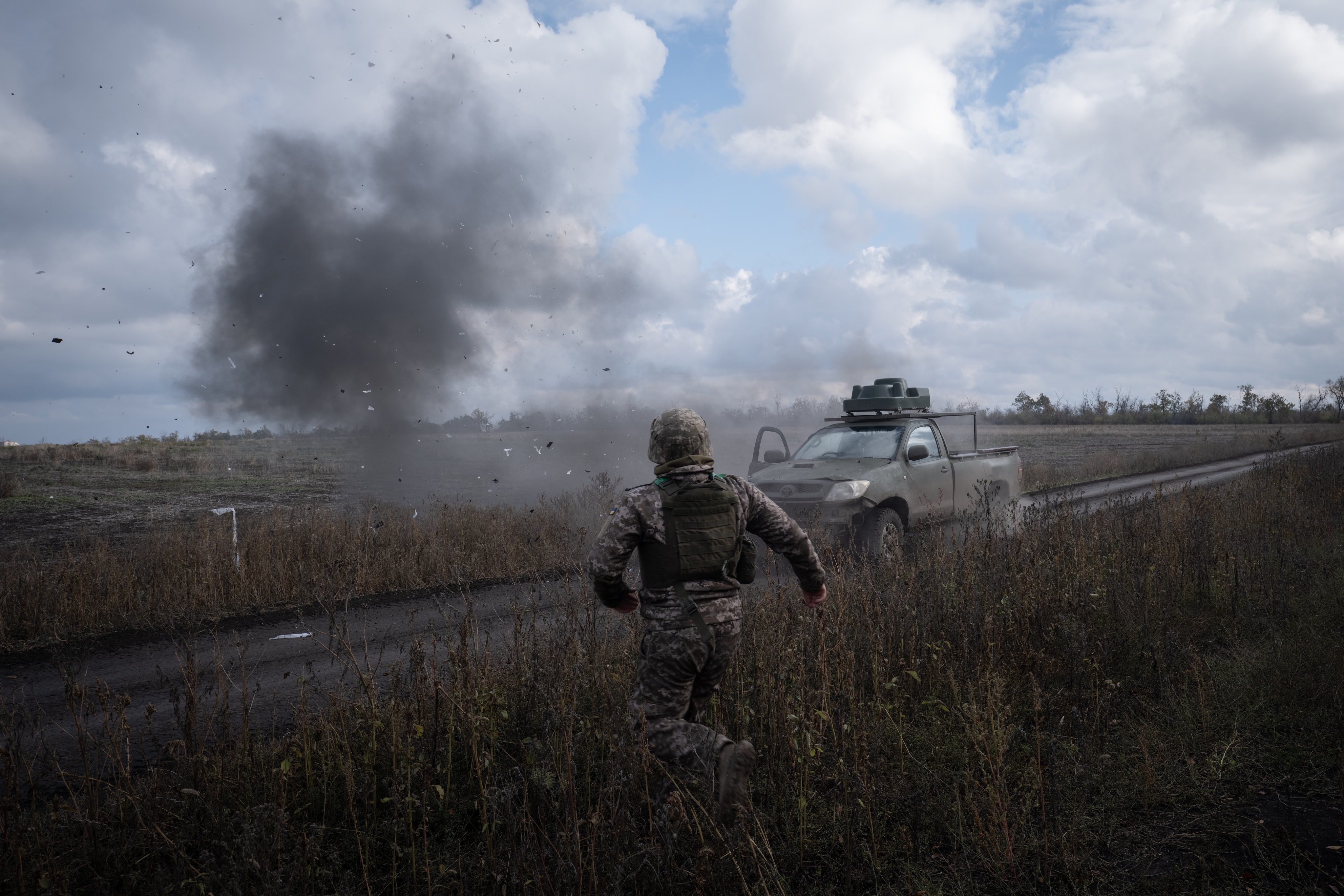 Soldier in camouflage and body armor runs along a dirt road as a plume of smoke and debris erupts beside a parked pickup truck in an open field.