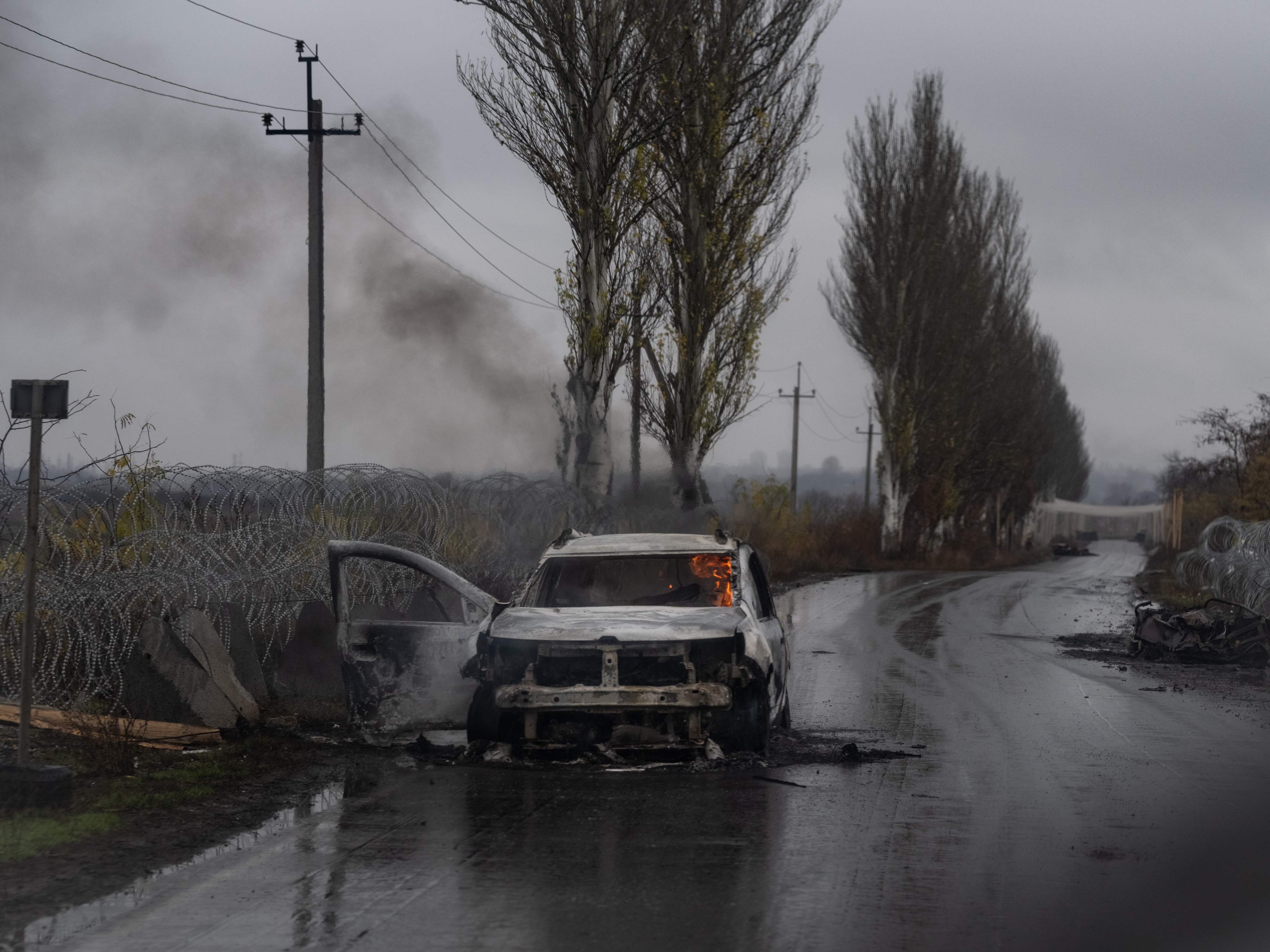 Burned-out car with driver door open and smoldering flames inside, abandoned on a wet road beside coils of barbed wire and tall poplar trees.