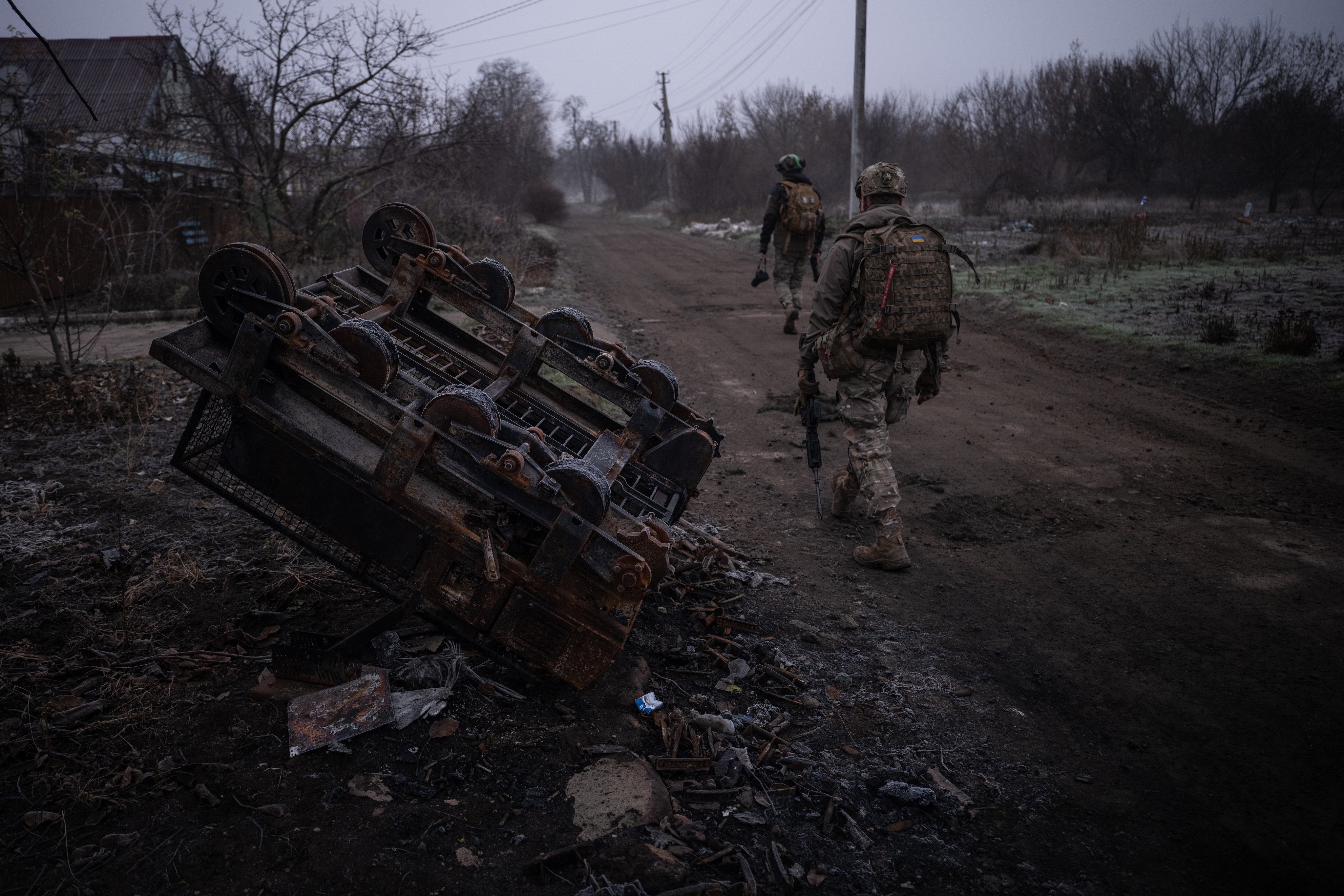 Two soldiers in camouflage with rifles and backpacks walk along a muddy road past an overturned rusted metal cart beside leafless trees.