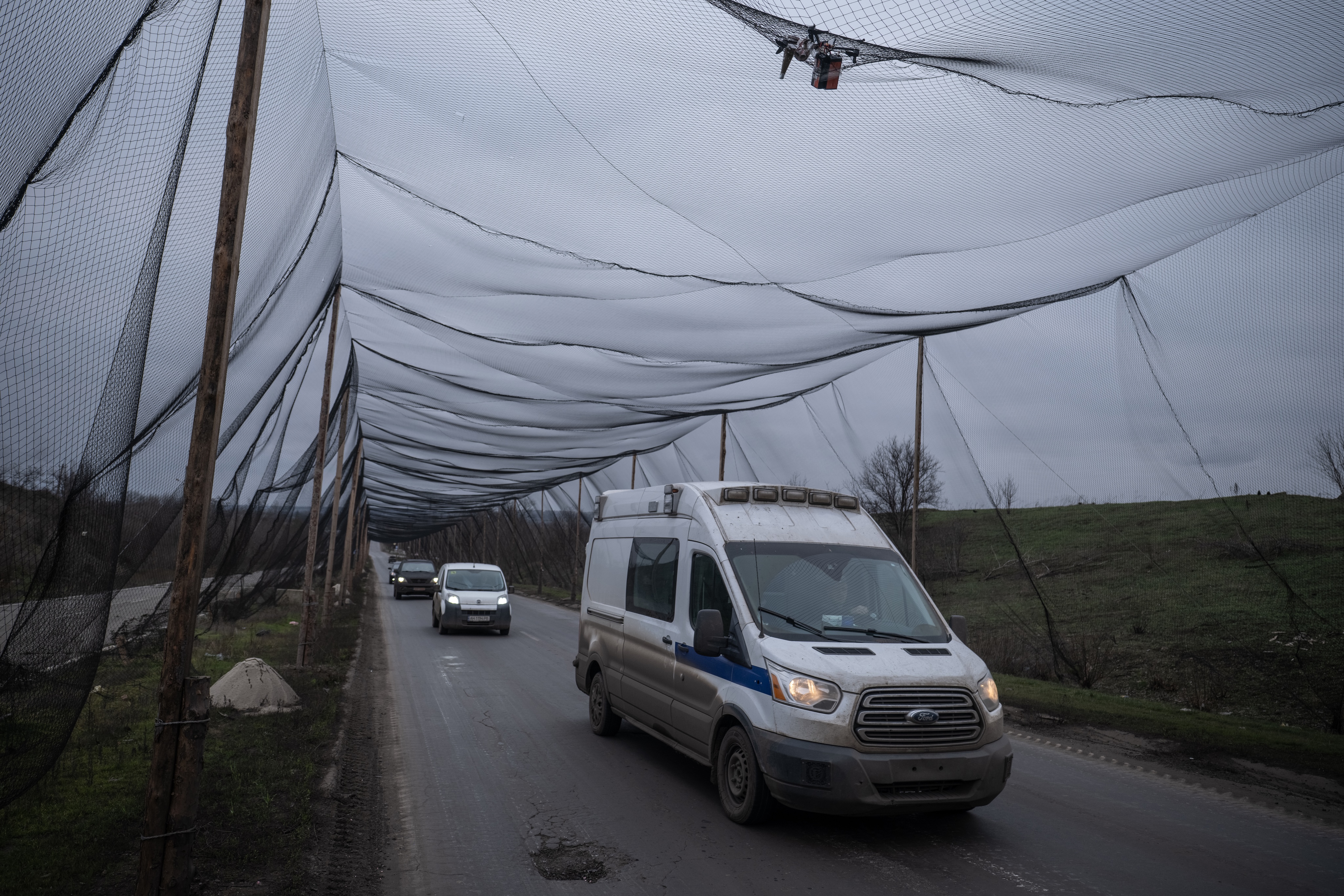 White van and other cars drive under large black mesh netting suspended on wooden poles over a rural road.