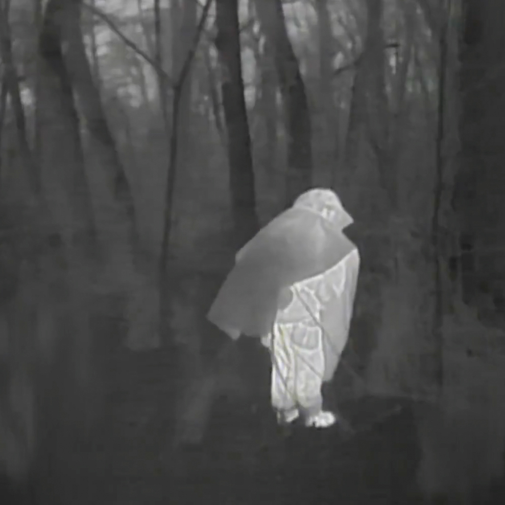 Person wrapped in a blanket or cape captured in a grainy night‑vision photo, standing among leafless trees in a dark forest.