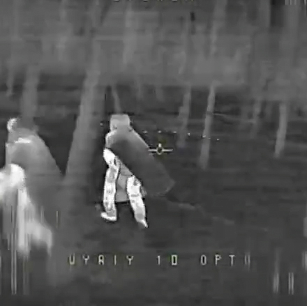 Thermal night-vision view of two people walking among trees; one carries a large cylindrical object on their shoulder.