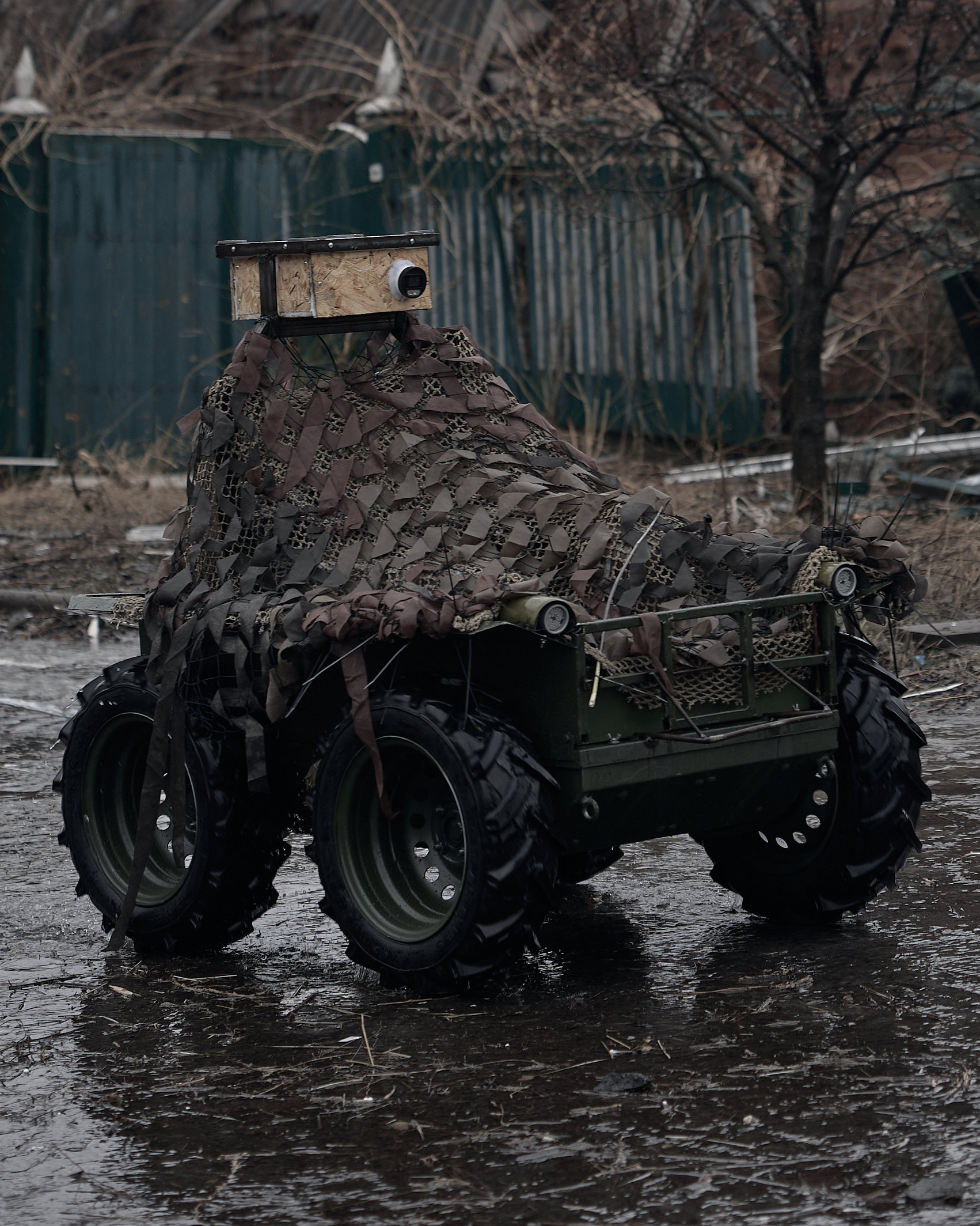 Small four-wheeled unmanned ground vehicle draped in camouflage netting, with a wooden box housing a camera-like sensor mounted on top.