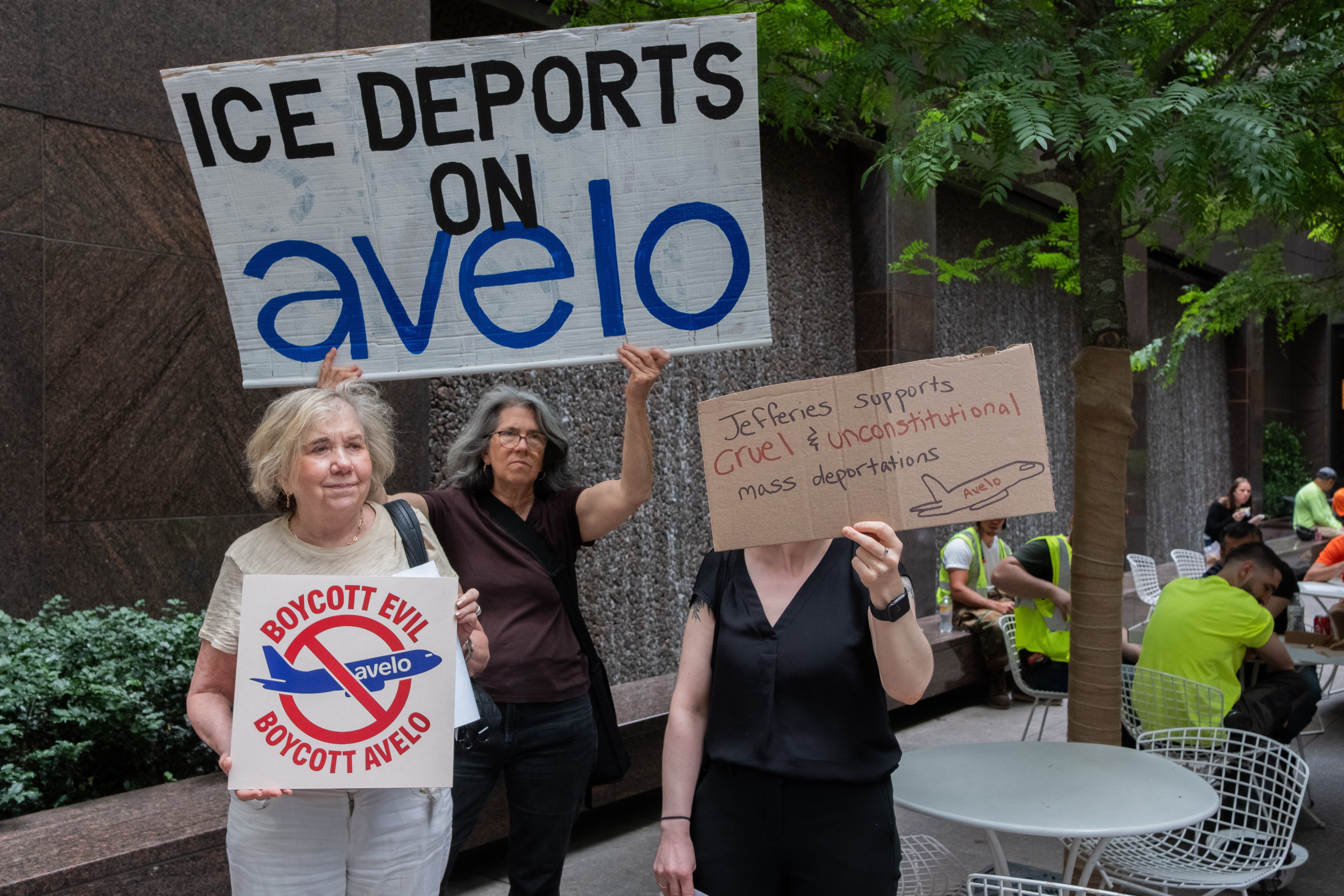 Three older women stand outside of an office building in Manhattan, holding cardboard signs reading “ICE Deports on Avelo”, “Boycott Evil, Boycott Avelo”, and “Jefferies supports cruel & unusual mass deportations.”