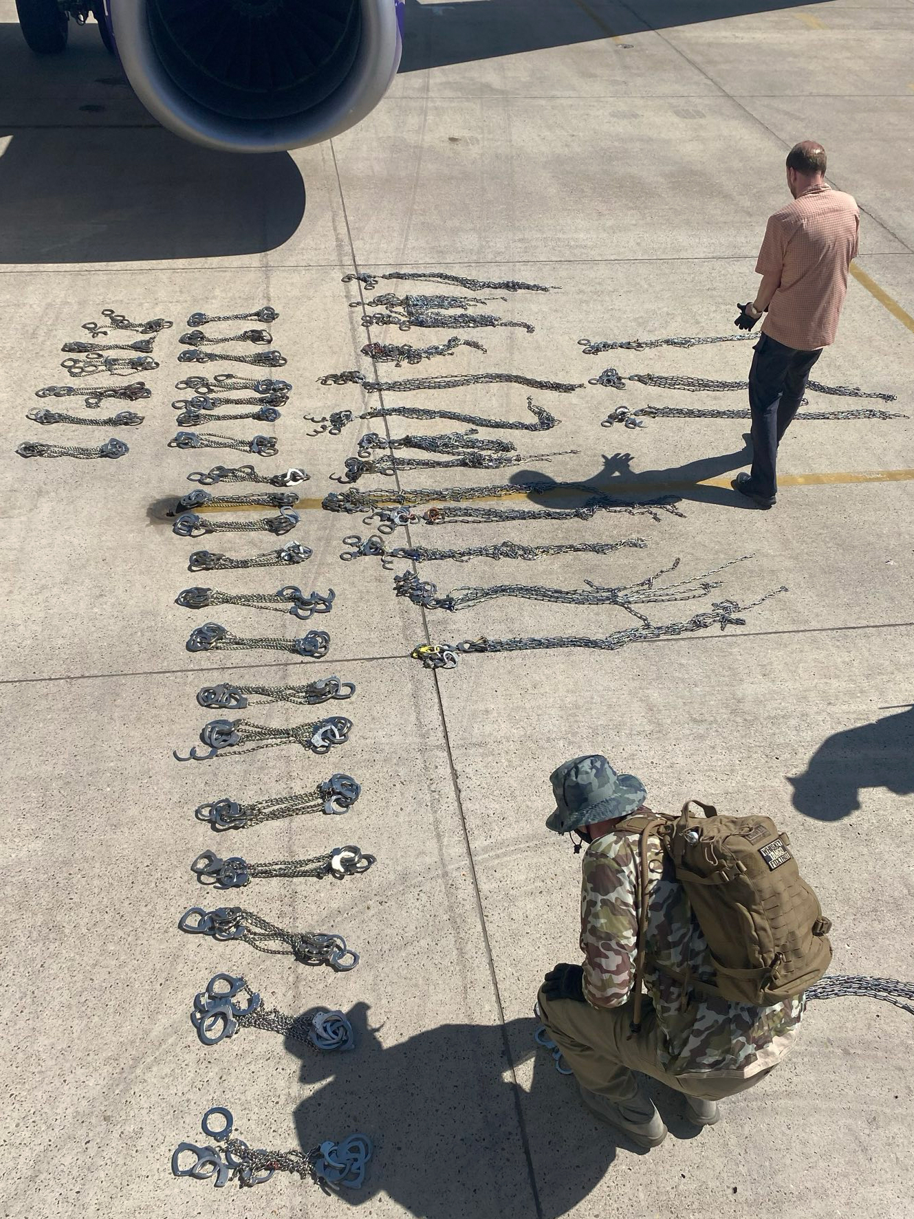 Two men lay sets of handcuffs and leg chains on the ground, counting them, just under the wing of an airplane.