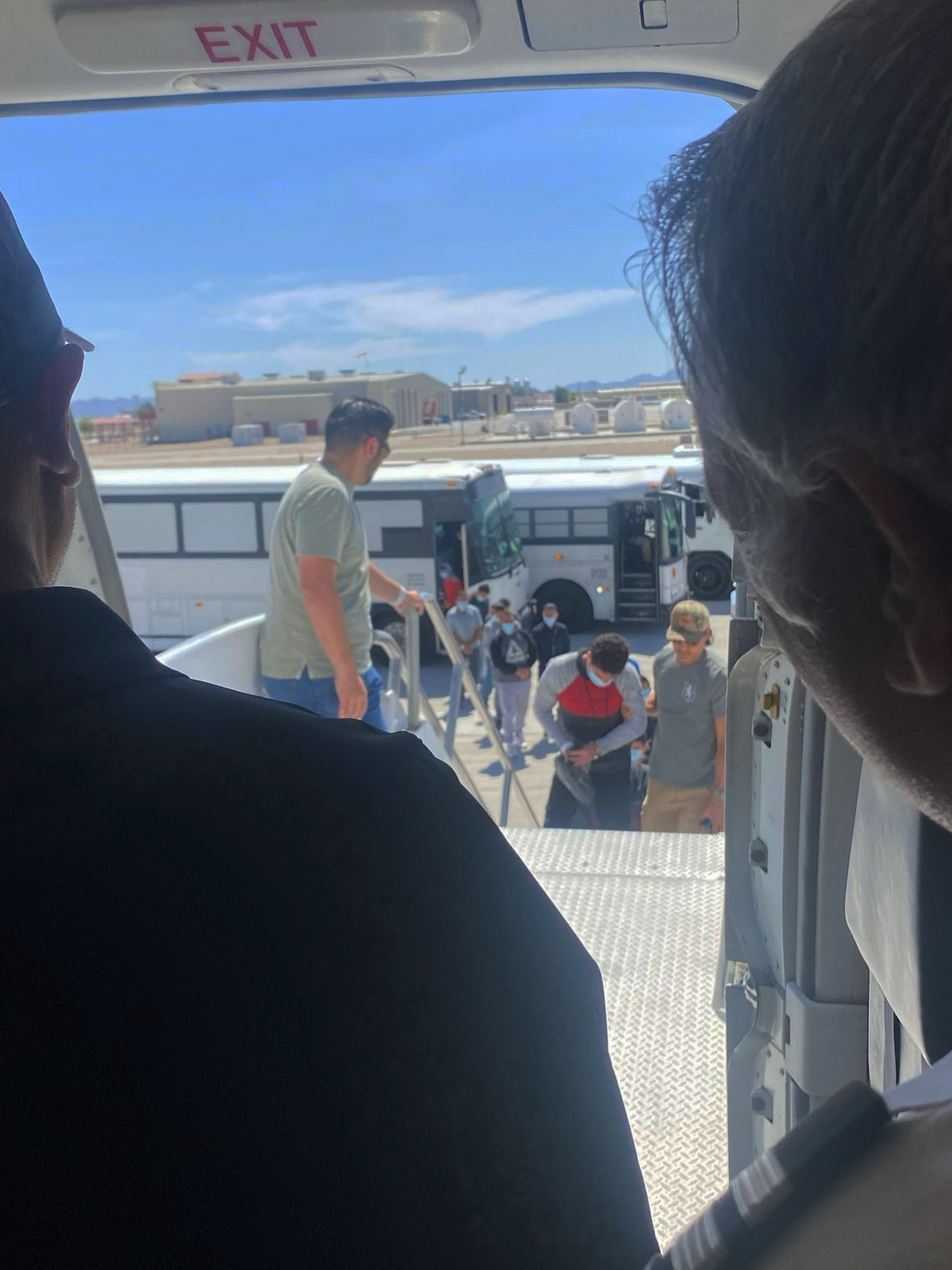 A view looking over the shoulder of an aeroplane pilot, out the main entrance door. Coming up the stairs to the door are two security guards escorting a detainee wearing chains and a blue surgical mask.