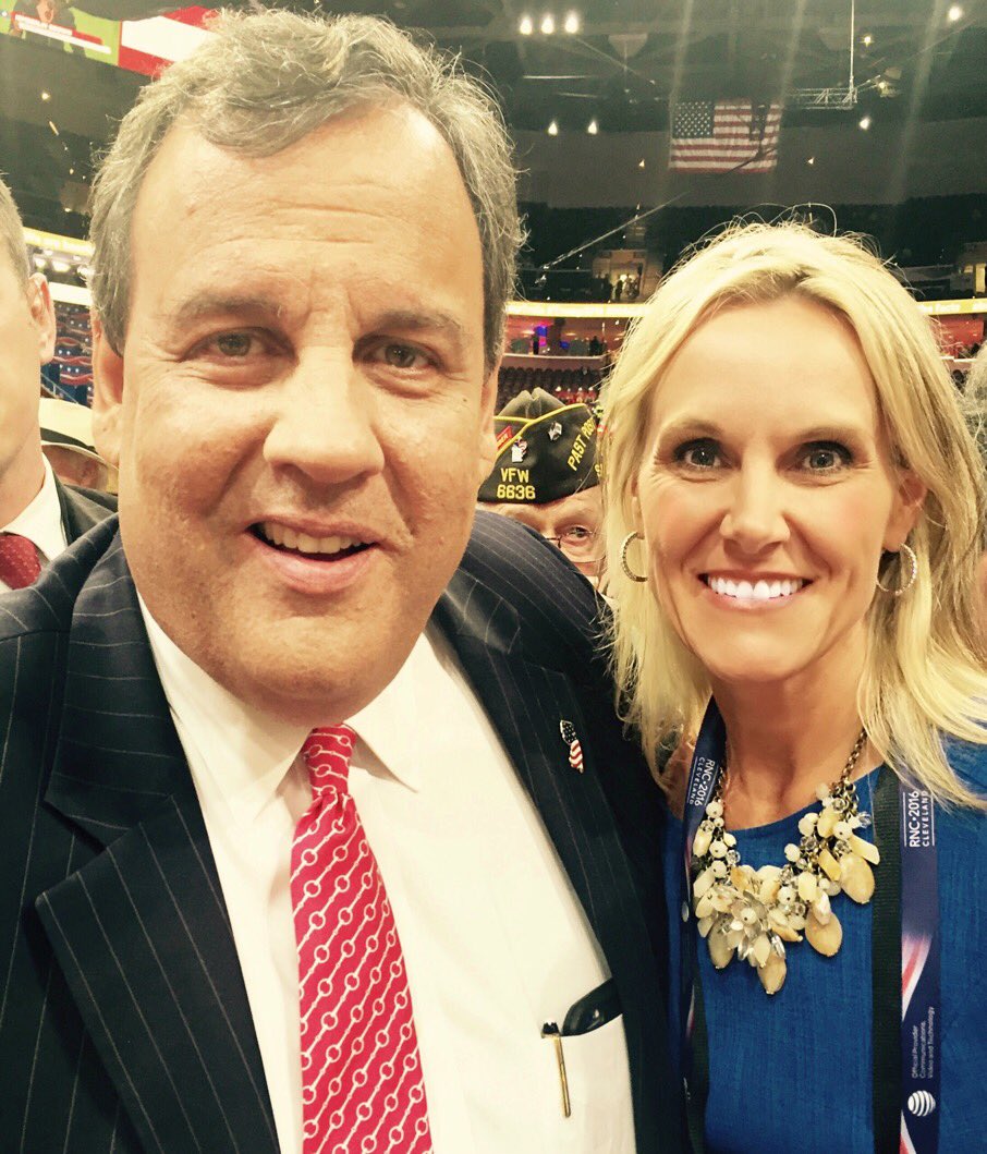 A selfie showing a smiling woman in blue standing next to a man in a suit and red tie. They stand in the crowded floor of a large arena.