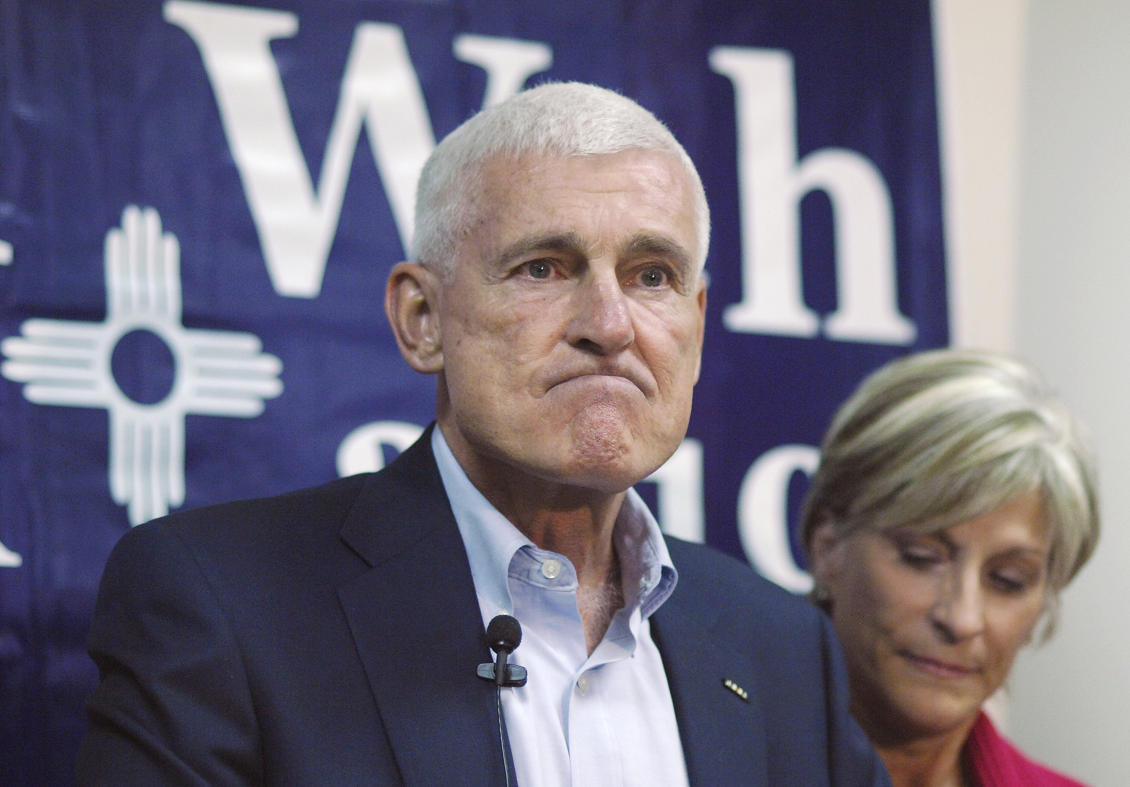 A man in a suit, frowning, in front of a sign that says “Allen Weh”.
