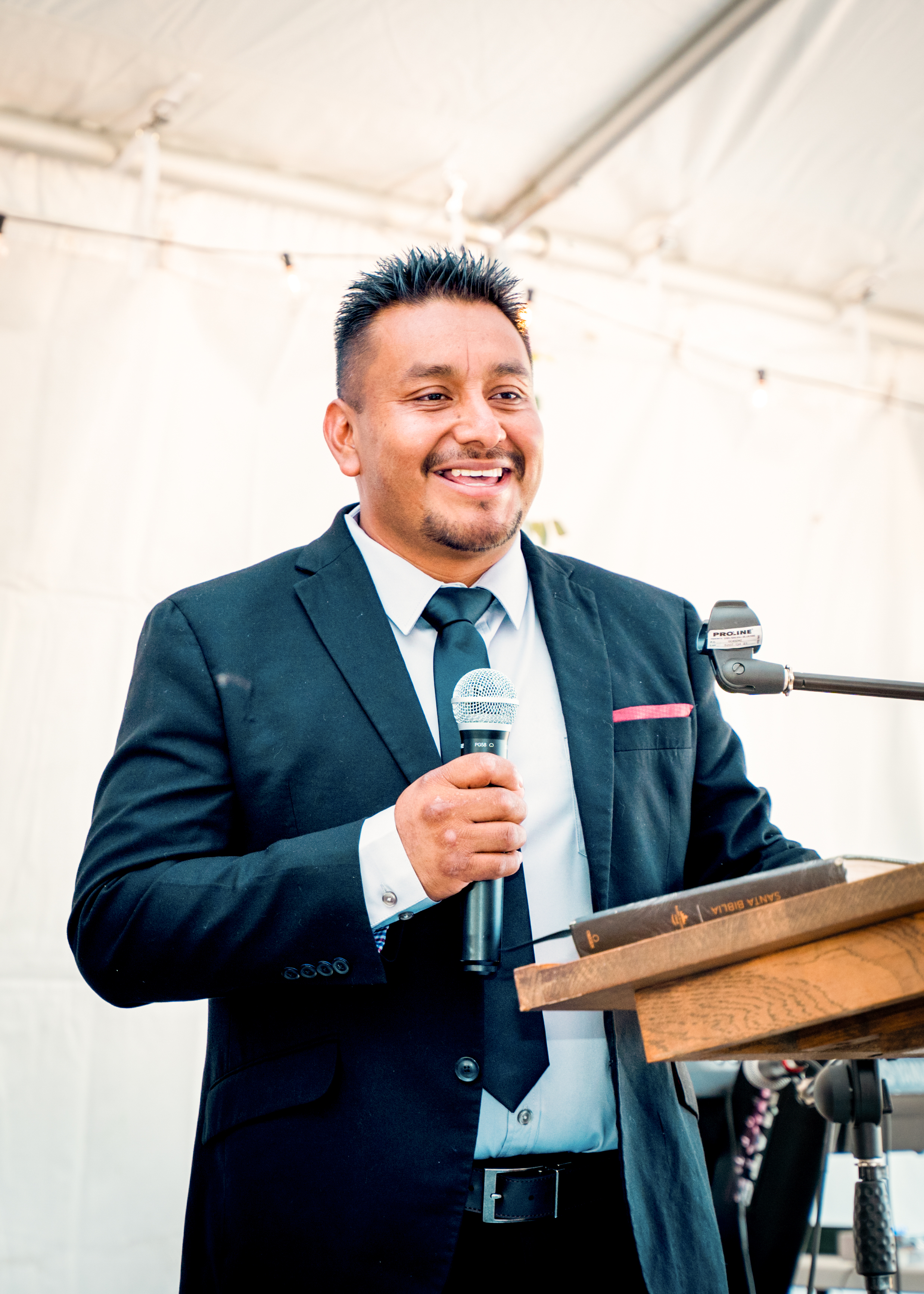 Maurilio Ambrocio, dressed in a suit and tie, smiles while standing at a podium holding a microphone at an event held in a white tent.