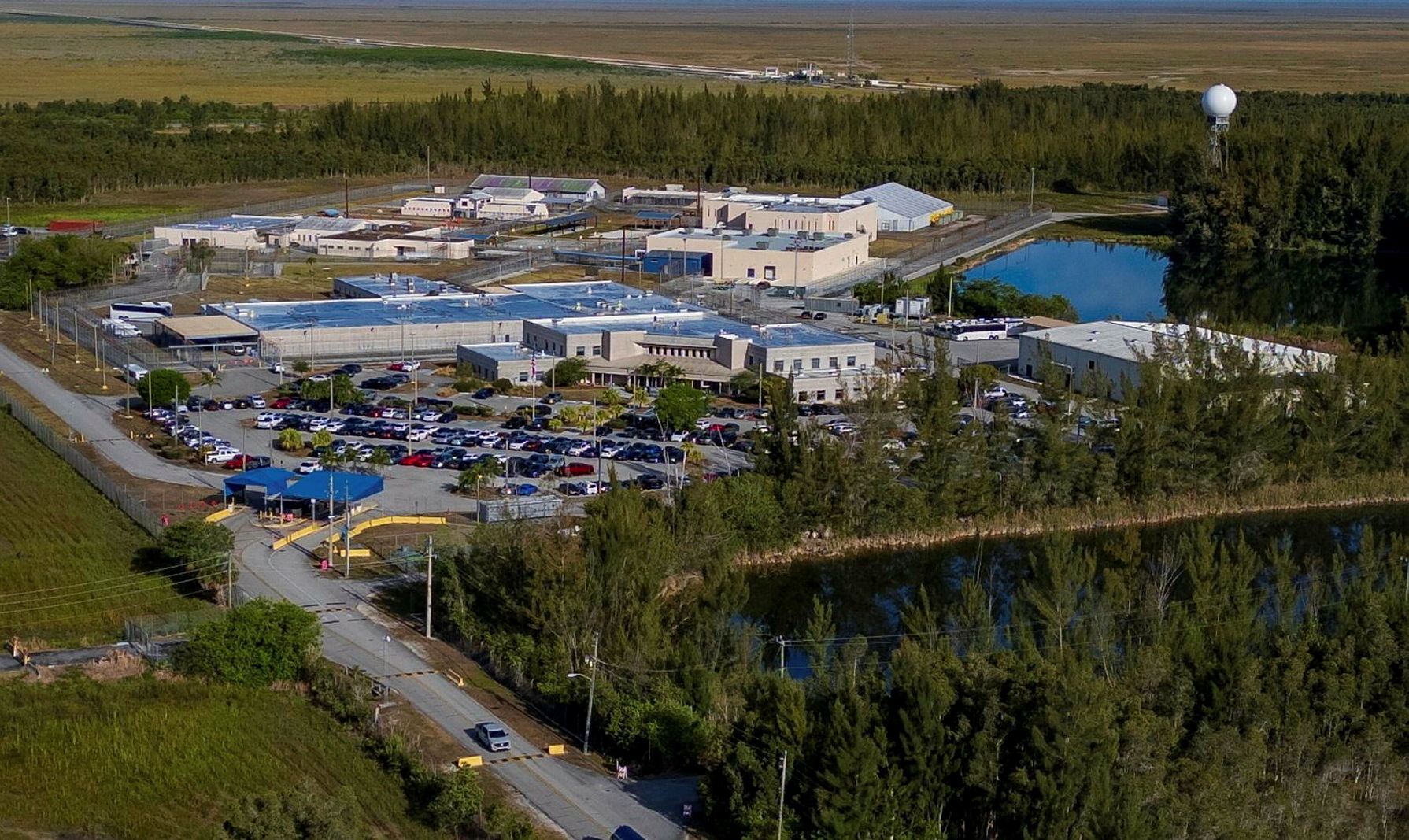 Aerial view of a large immigration detention facility surrounded by fencing, with multiple beige and blue buildings, a crowded parking lot, and a gated entrance, set near a wooded area and water.