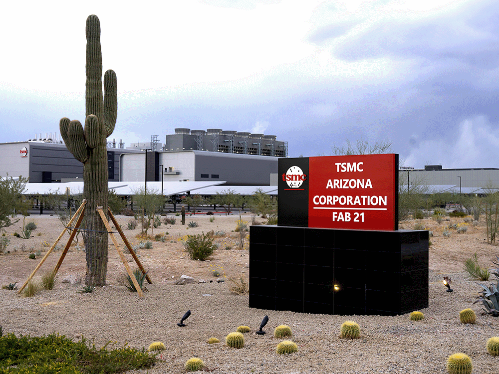 Ground-level image of the TSMC Arizona Corporation Fab 21 facility in Phoenix, Arizona. A large red and black sign with the TSMC logo stands in the foreground, with a desert landscape featuring cacti and gravel. The modern industrial building is visible in the background under a cloudy sky.