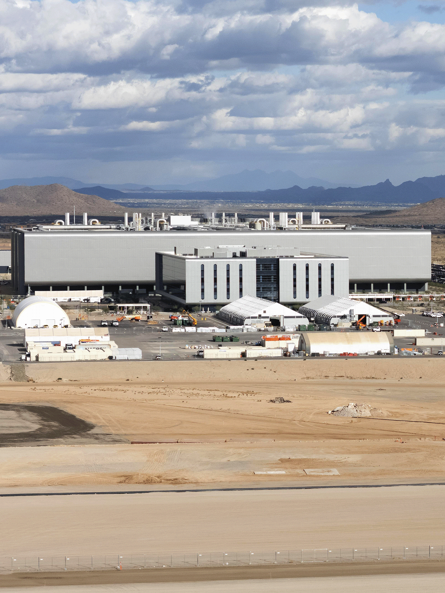Distant view of the TSMC facility in Phoenix, Arizona. The large, modern building is set against a backdrop of desert mountains and dramatic cloudy skies. Construction equipment, modular buildings and temporary structures are visible in the foreground, along with a wide expanse of cleared desert land.