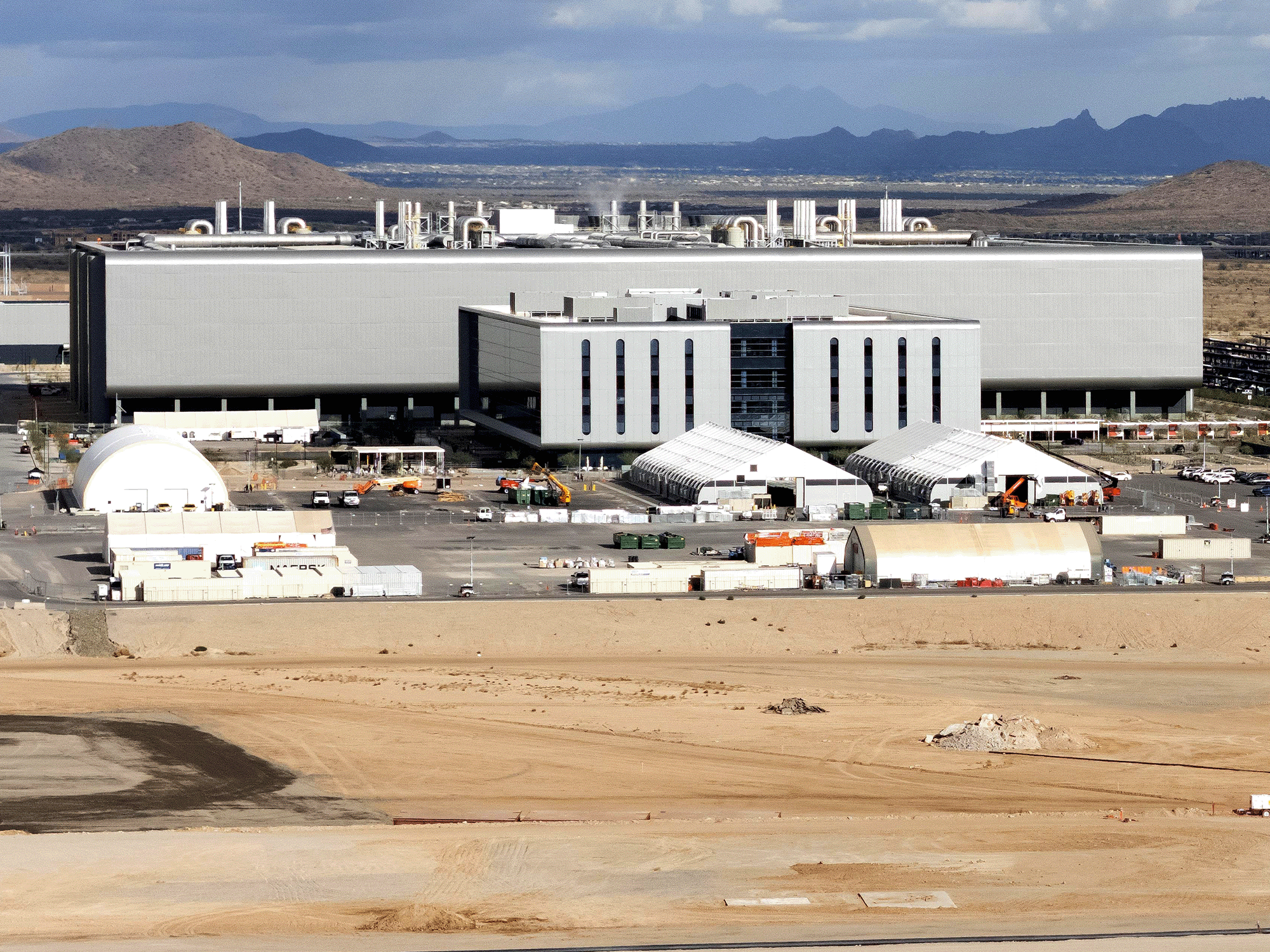 Distant view of the TSMC facility in Phoenix, Arizona. The large, modern building is set against a backdrop of desert mountains and dramatic cloudy skies. Construction equipment, modular buildings and temporary structures are visible in the foreground, along with a wide expanse of cleared desert land.