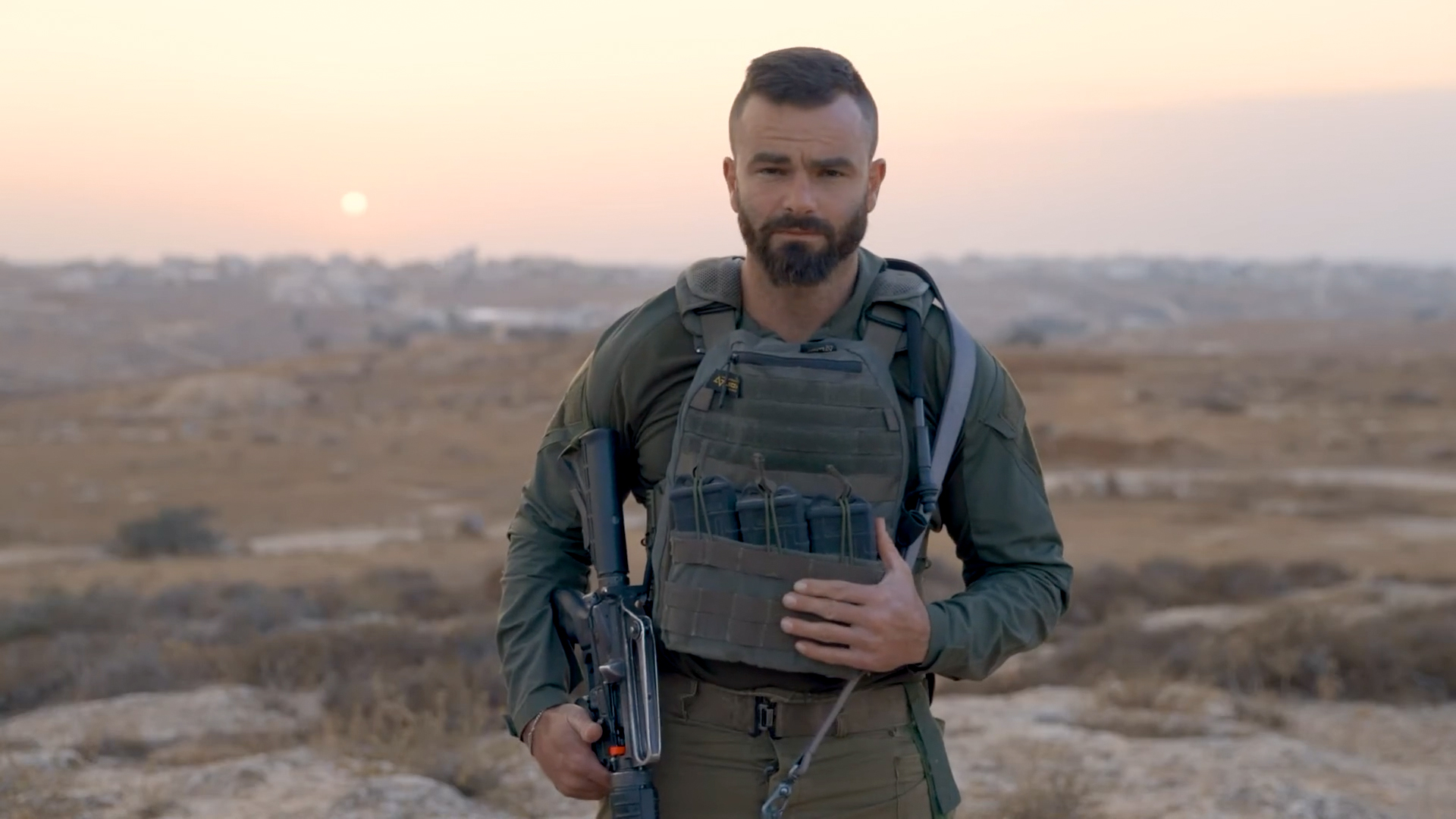 A still from a video showing a man in olive-green military gear holding an assault rifle to his side. His hair is cropped short at the sides and he has a beard. Behind him is a rocky landscape with a setting sun.