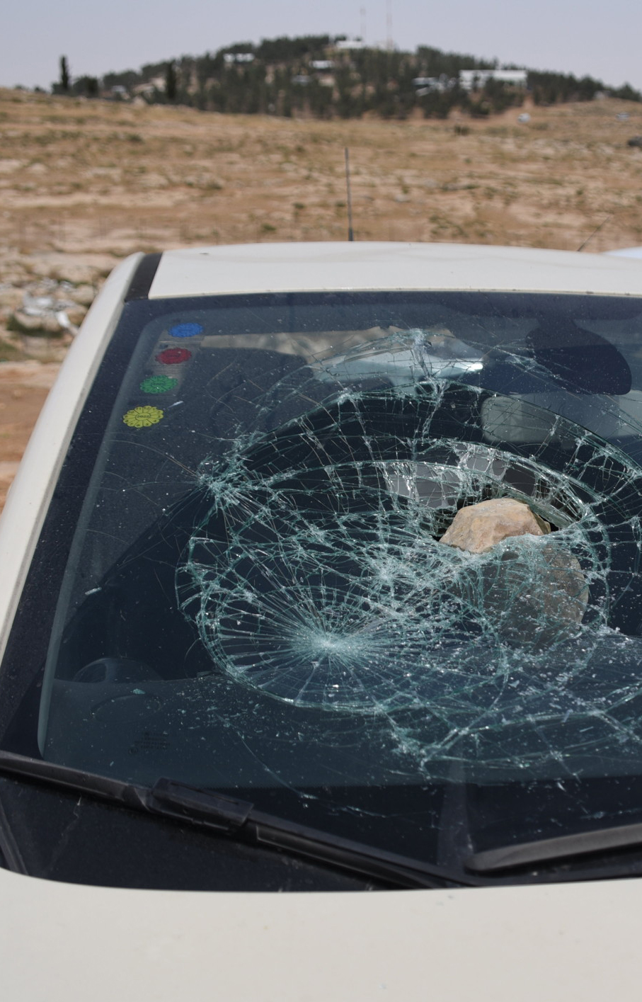 A white car with a large rock smashed through the windscreen. In the background is a hill with trees and buildings.