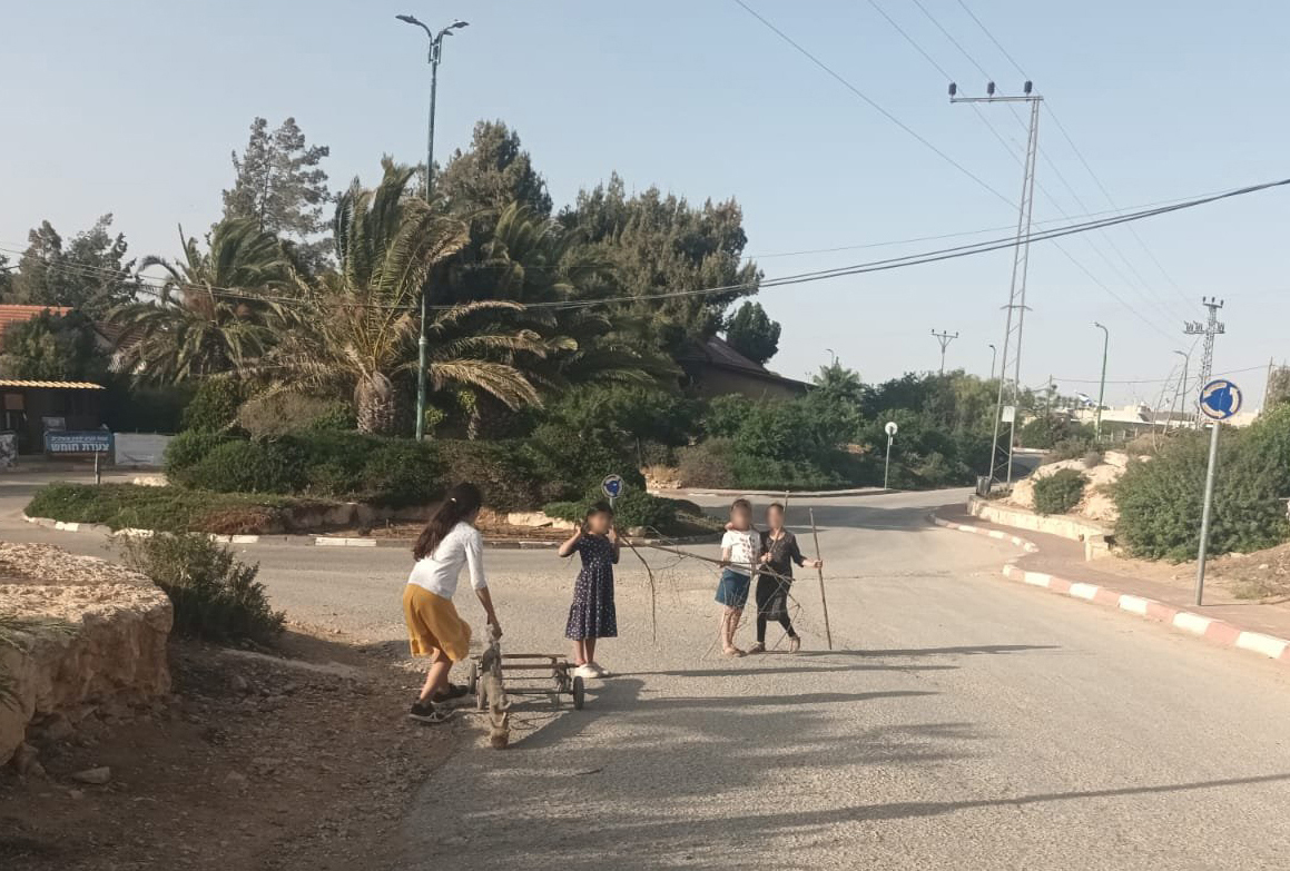 A street in Ma’on. In the foreground, four young girls play with sticks. In the background, there is a roundabout with green hedges and a large palm tree. Power lines and vegetation continue into the distance.