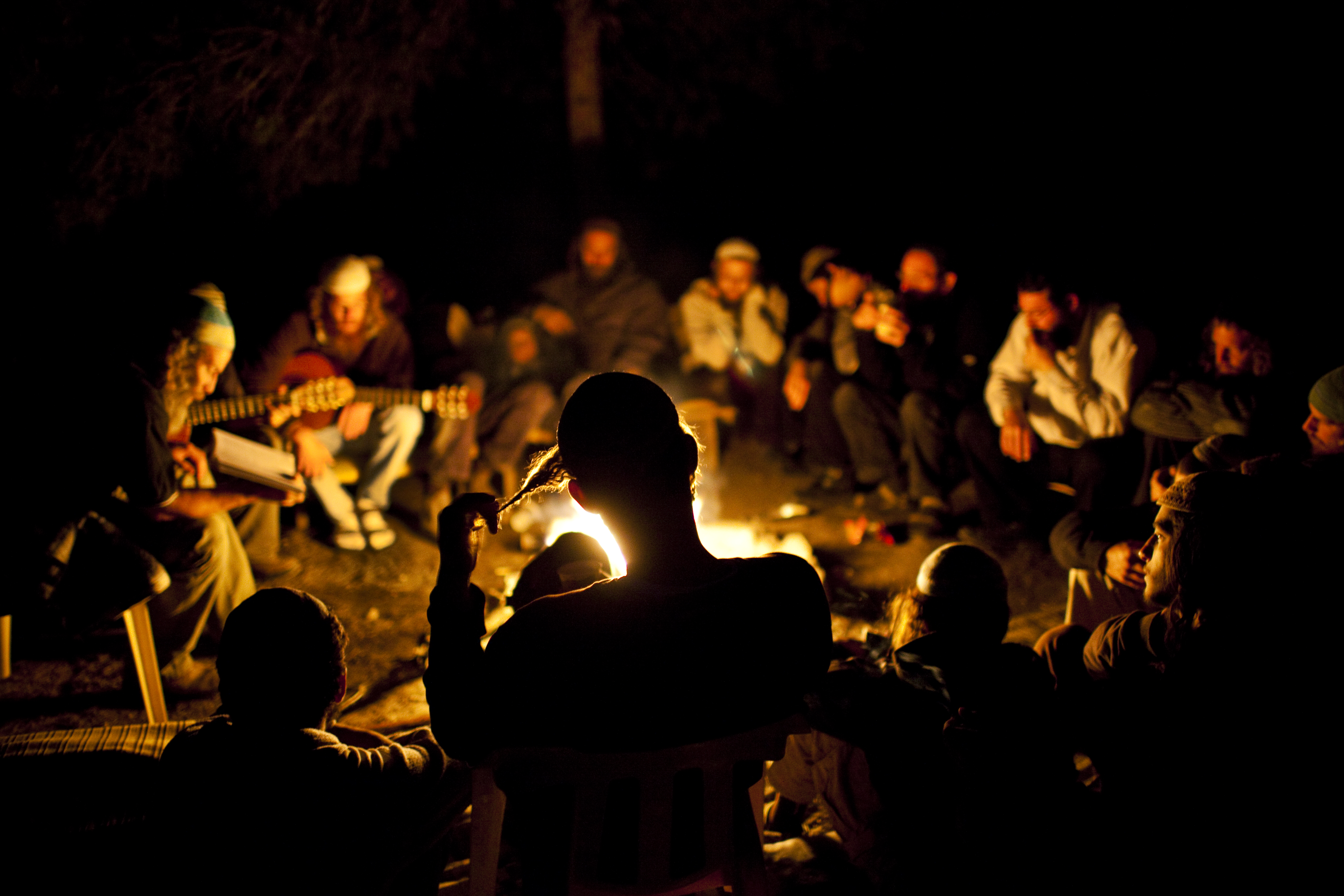 A group of men sitting around a campfire at night. Some men have guitars. One man is silhouetted in the foreground pulling on his payot.