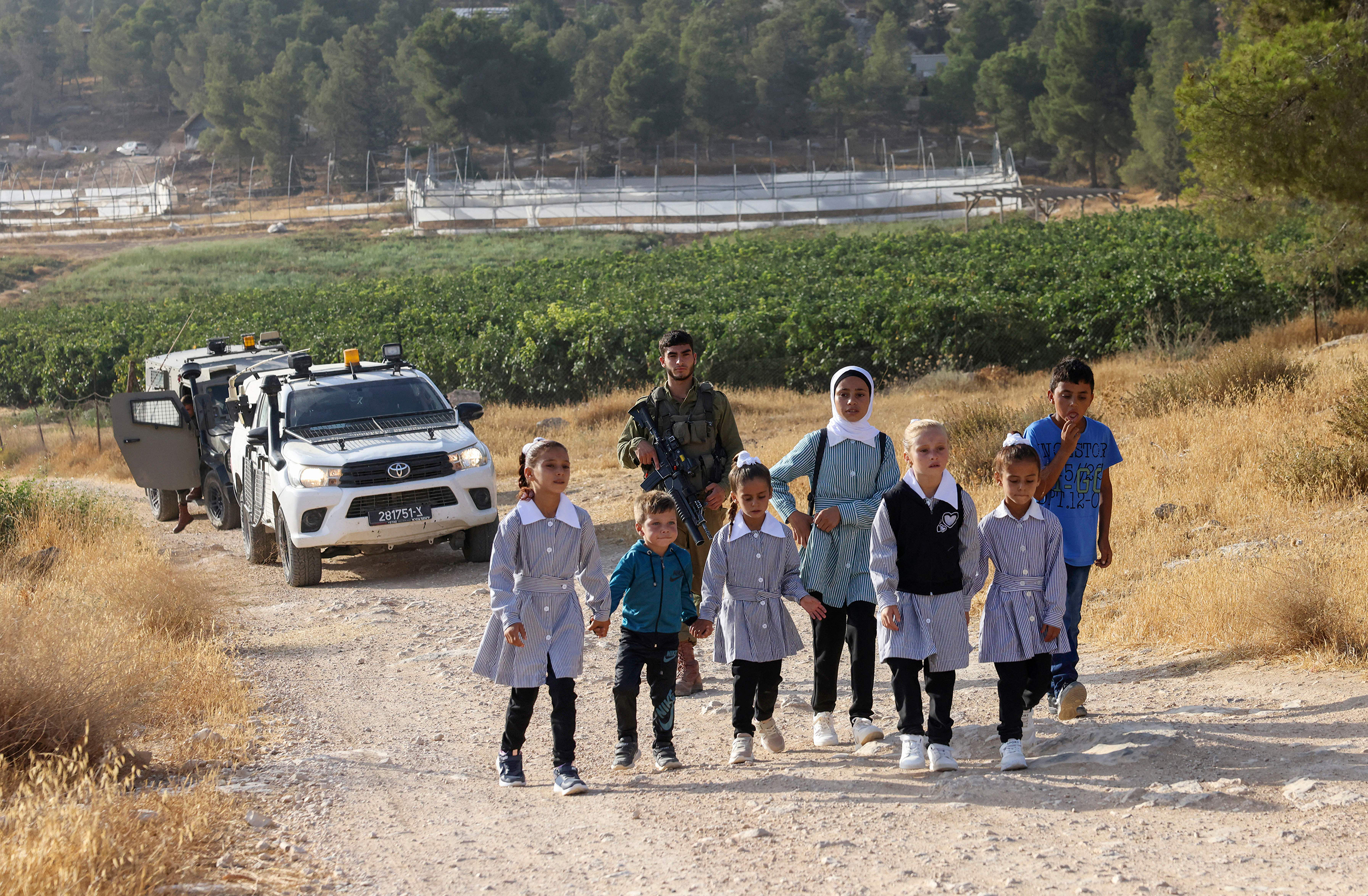 A group of seven schoolchildren are walking down a road, accompanied by a soldier with a rifle. In the background, agricultural fields and trees are visible as well as military vehicles.