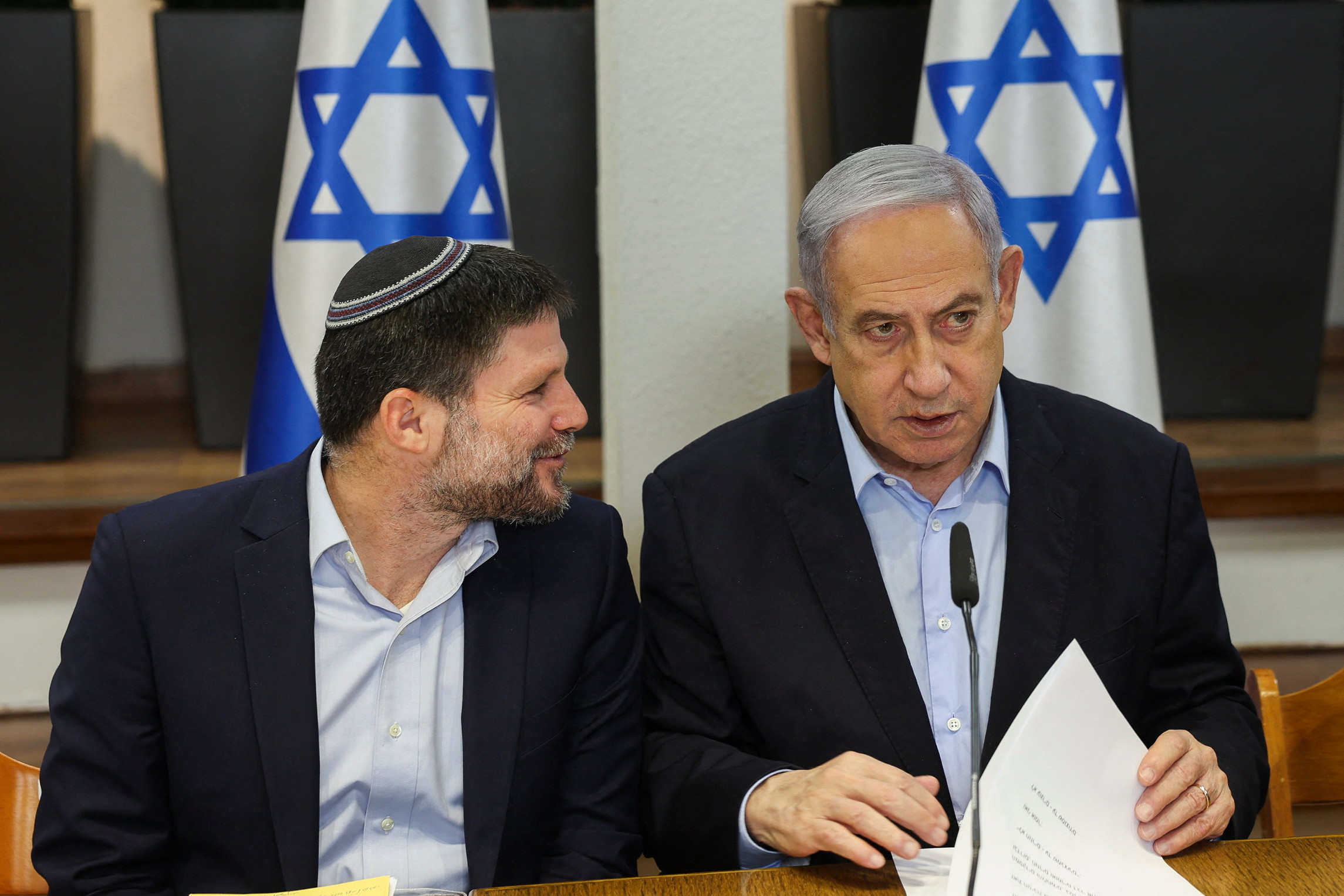 Two men in dark suit jackets and light blue shirts sit at a desk in front of two Israeli flags. The man on the left is wearing a kippah and looking at the man on the right.