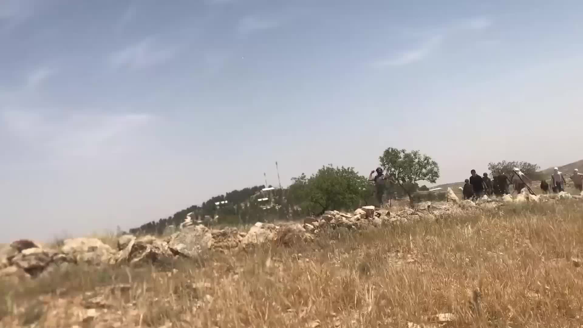 A video showing a group of men standing on a verge in a hilly landscape. They spot the person filming and begin to run towards the camera, some throwing stones. The cameraman retreats, shaking the camera.